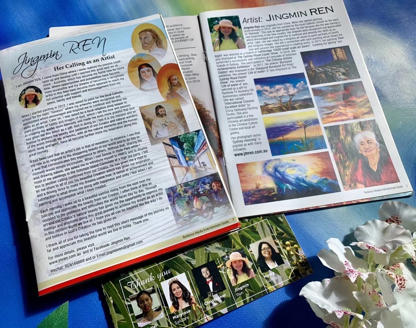 Open magazine features a biography of artist Jingmin Ren with her photos and artwork samples, alongside a page showcasing her paintings and achievements. The magazine is on a blue surface with white orchids nearby, and a thank you card with photos of