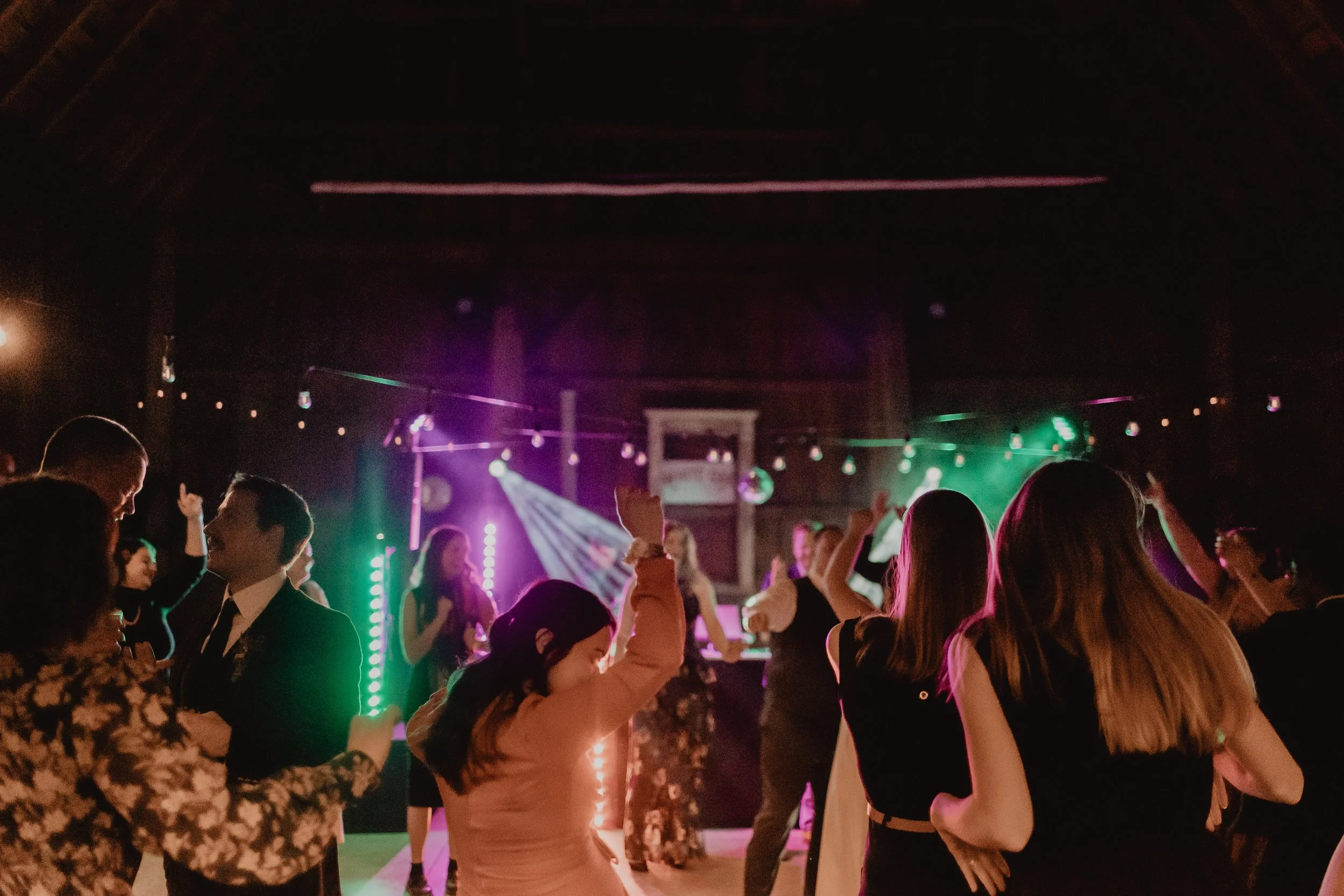 People dancing and socializing at a party with colorful lights and a DJ setup in the background.