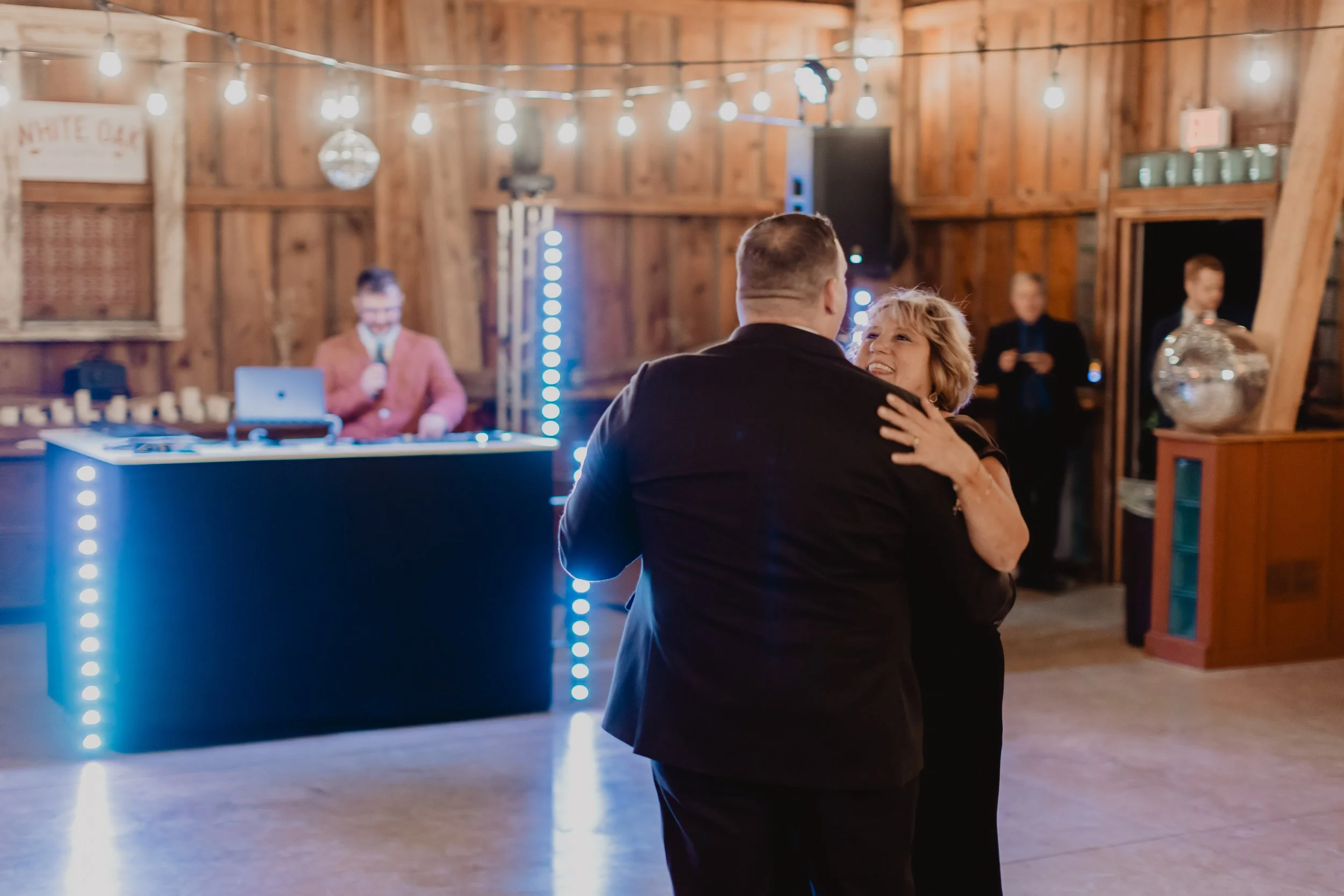 A couple dancing in a rustic barn at an event with string lights hanging overhead and a DJ on the stage in the background.
