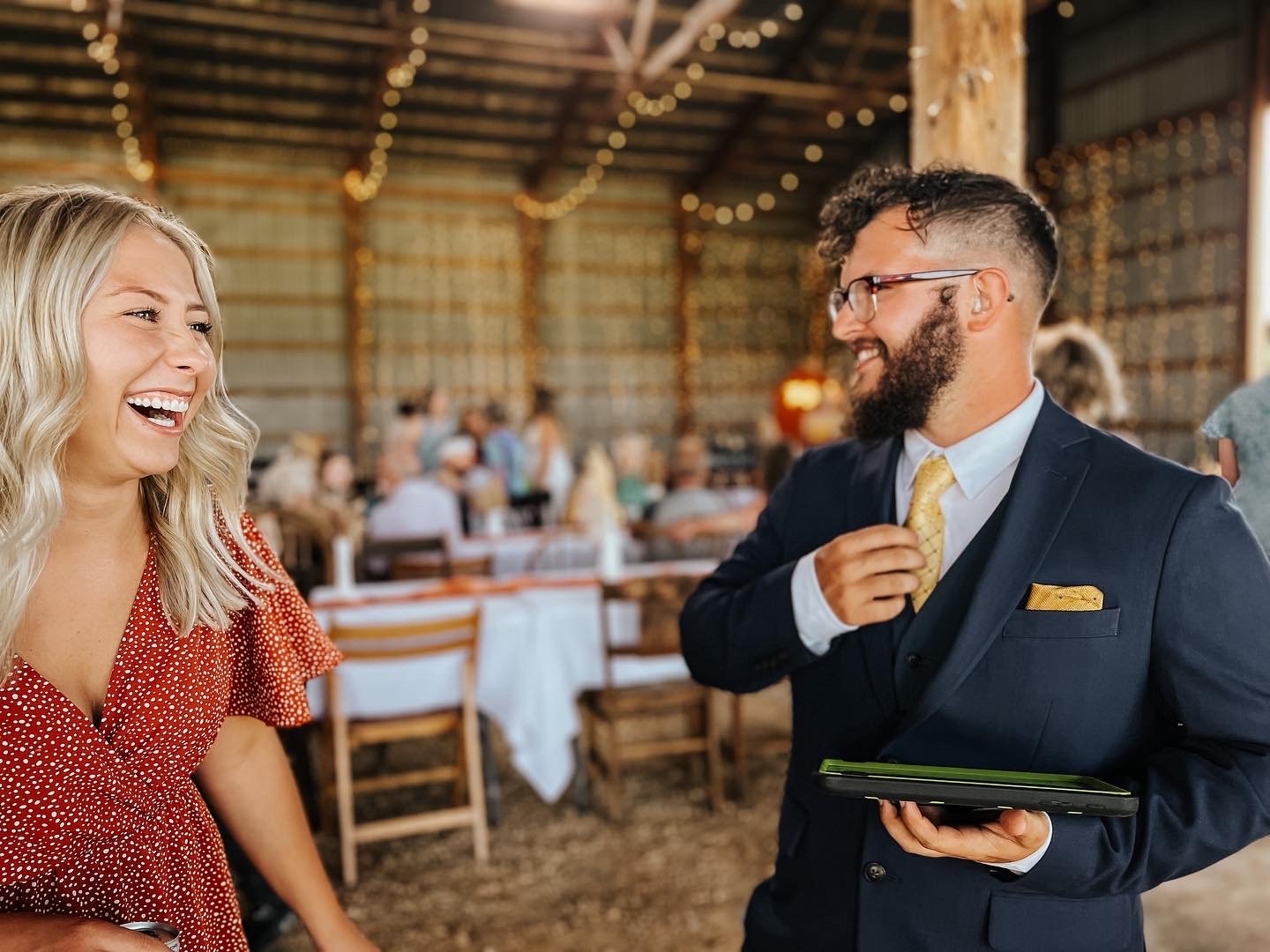 A man in a suit and a woman in a red dress laughing and smiling at each other, in a rustic barn decorated with string lights, at a social event or wedding reception.