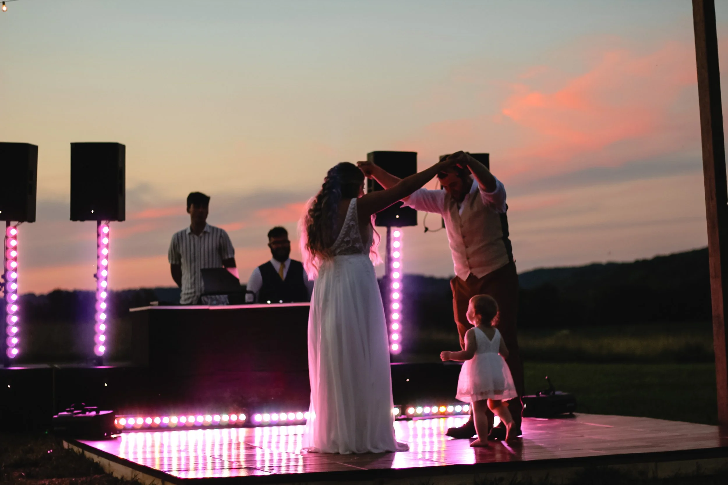 A couple dancing on a wooden platform outdoors at sunset with a young girl watching. The scene features colorful lighting and a DJ booth in the background.