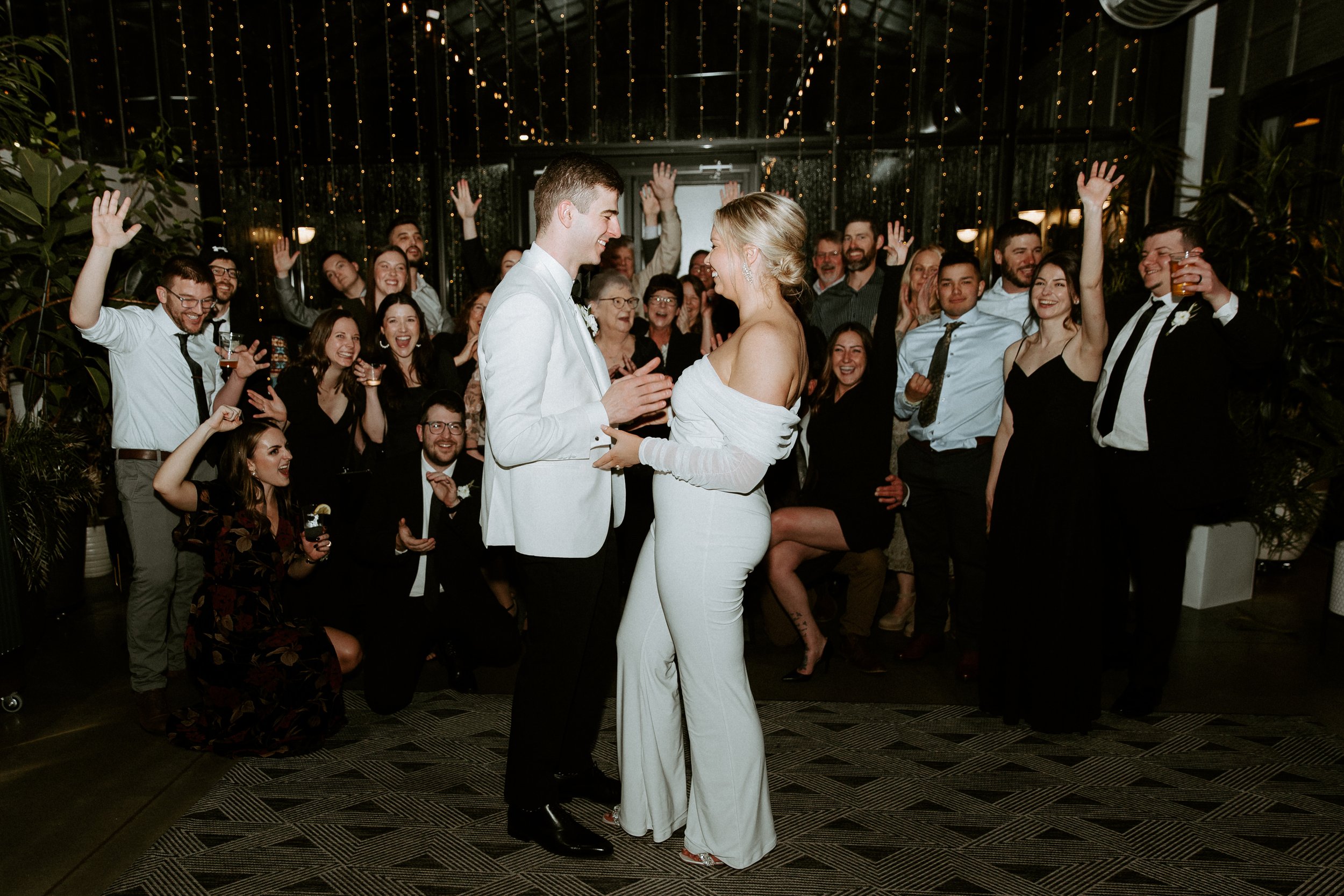 A bride and groom sharing a dance at their wedding reception, surrounded by smiling guests with raised hands, in a decorated indoor venue with string lights.
