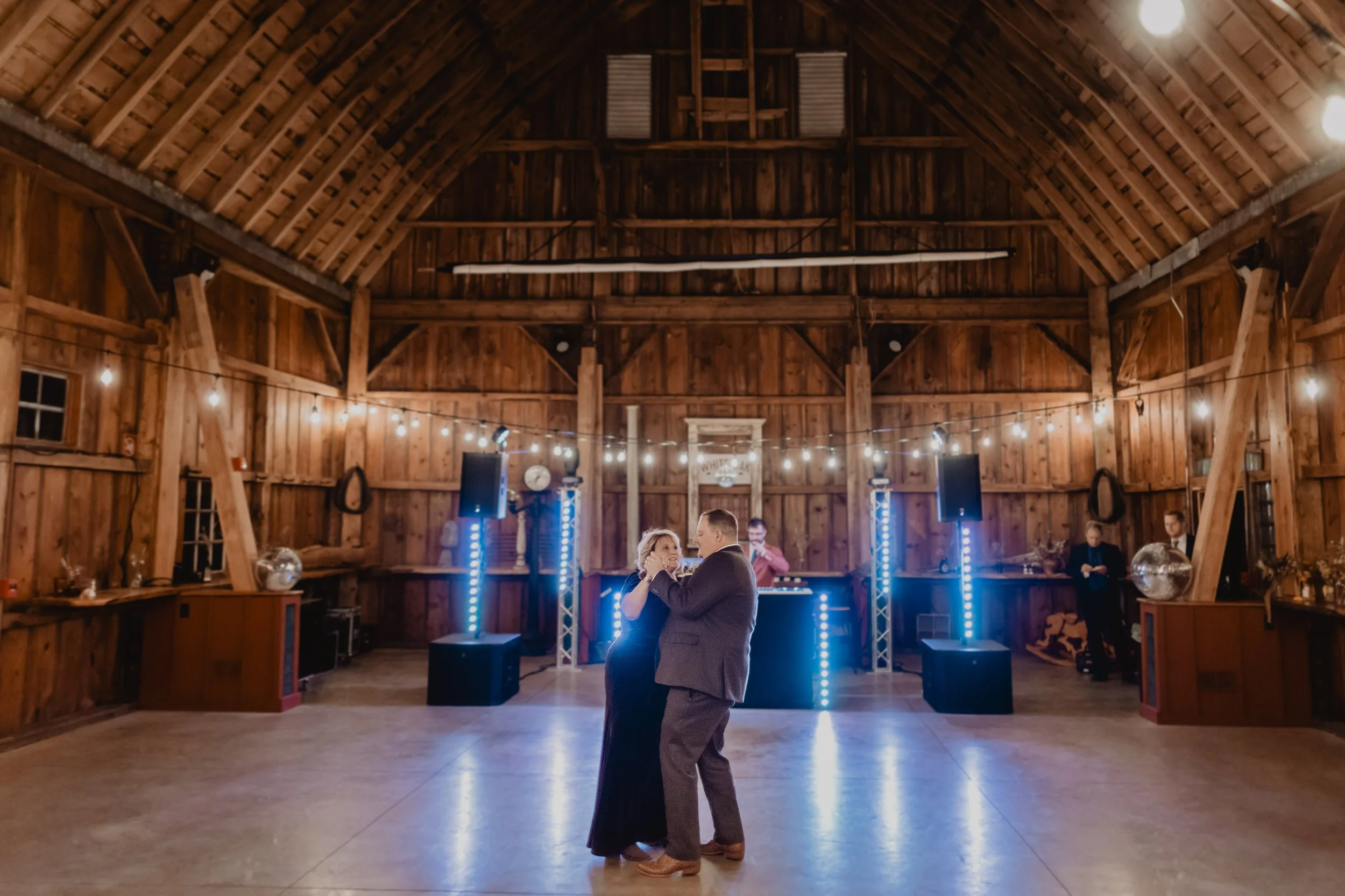 A couple dancing in a barn decorated with string lights, with a DJ setup and a few people watching in the background.