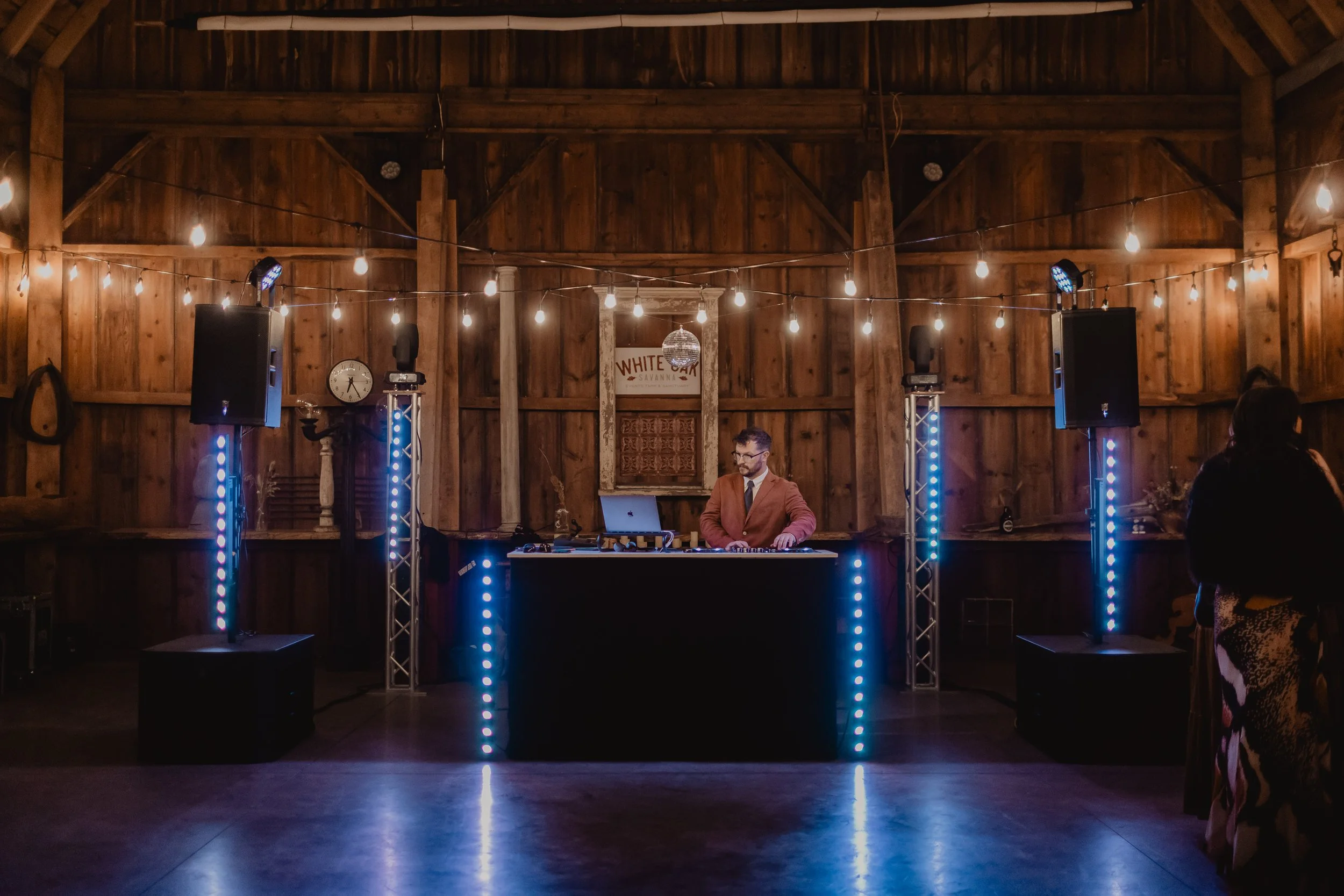 A man in a brown jacket and glasses is DJing at a wooden venue with string lights overhead. There are two large speakers on either side of him, and a small disco ball hanging from the ceiling. A sign with the words "WHITE OAK" is on the wall behind h