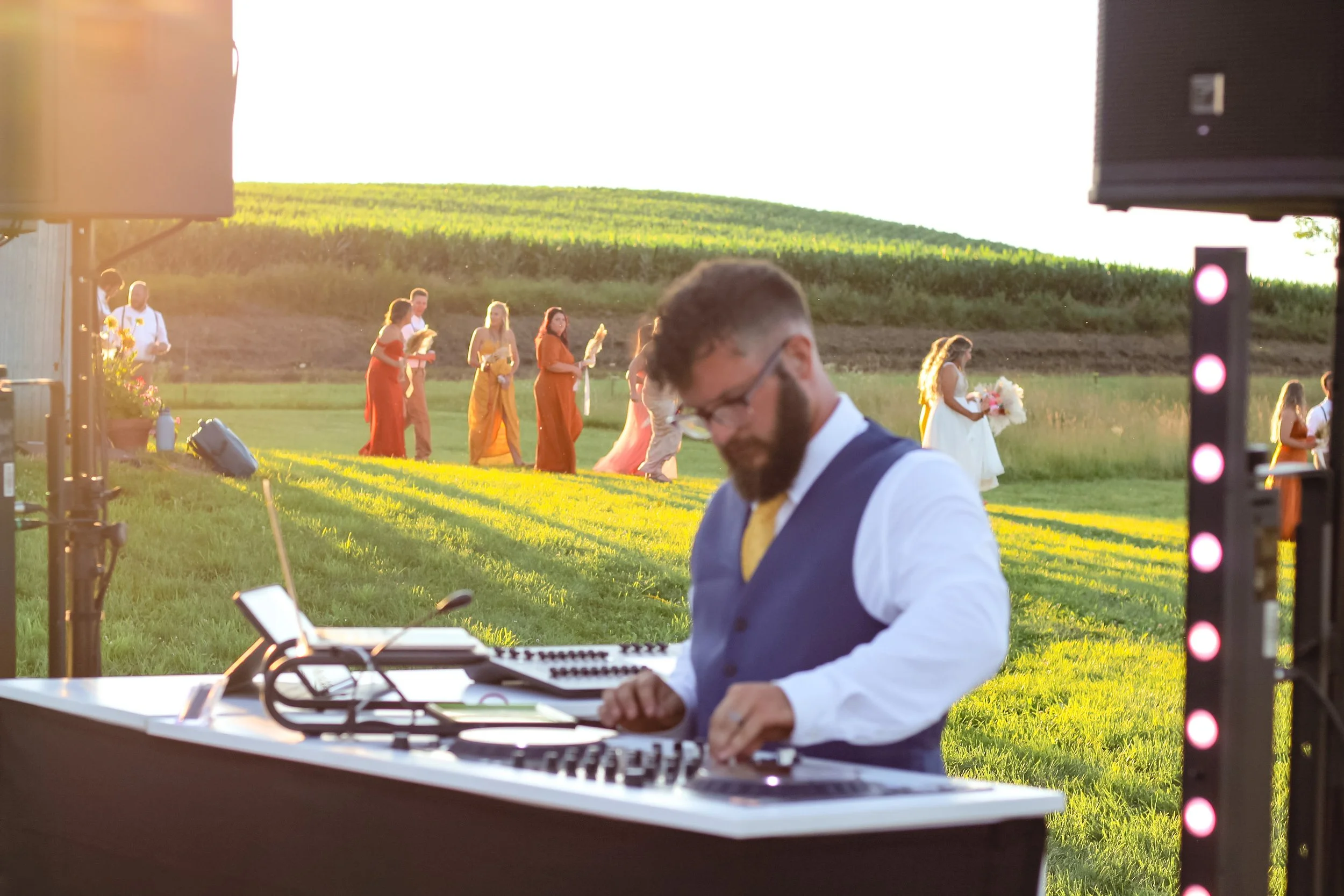 A DJ in a blue vest and white shirt plays music at an outdoor wedding reception during sunset, with the bride and bridesmaids in colorful dresses holding bouquets in the background on a lush green field.