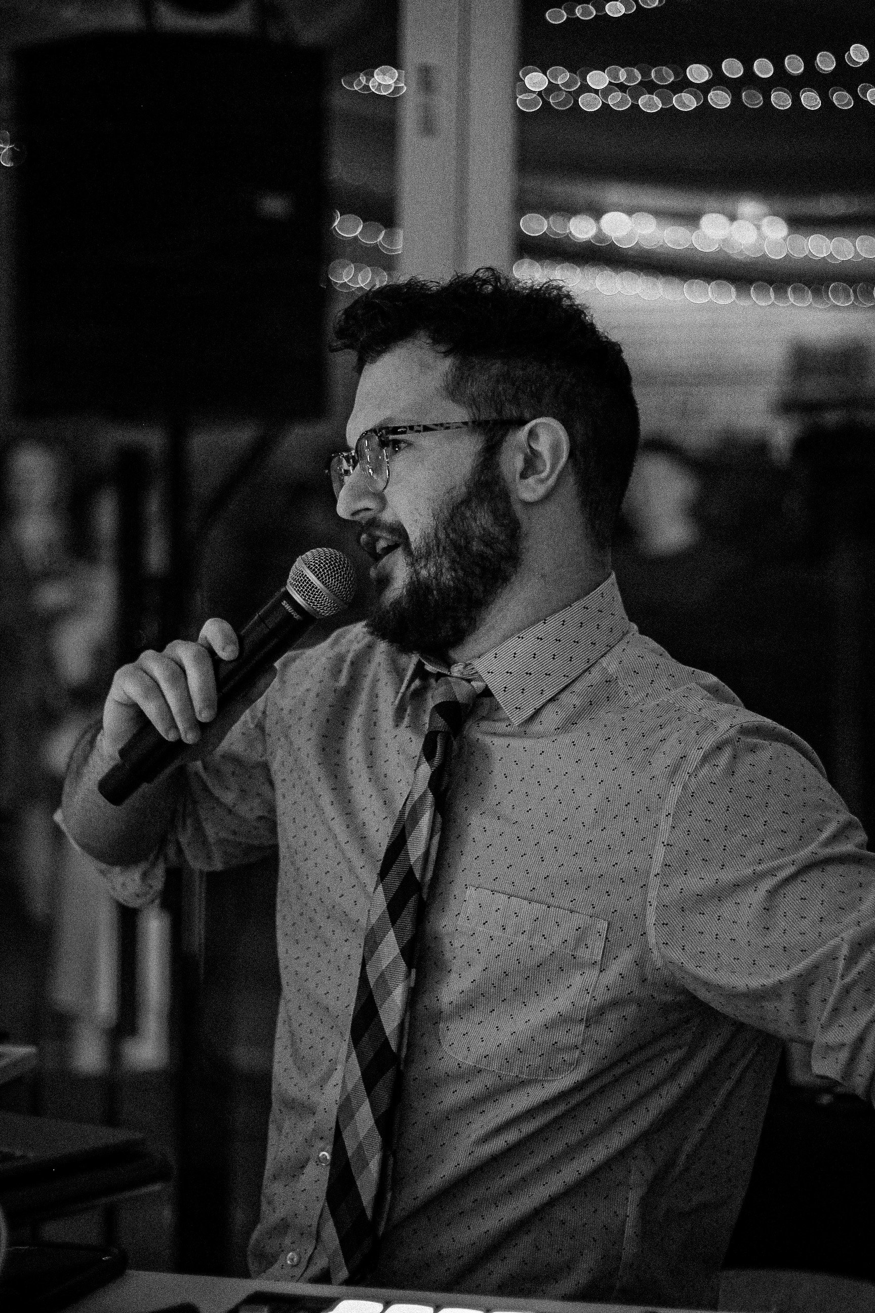 A man with glasses and a beard wearing a dotted shirt and a striped tie, holding a microphone near his mouth, appears to be speaking at an event.