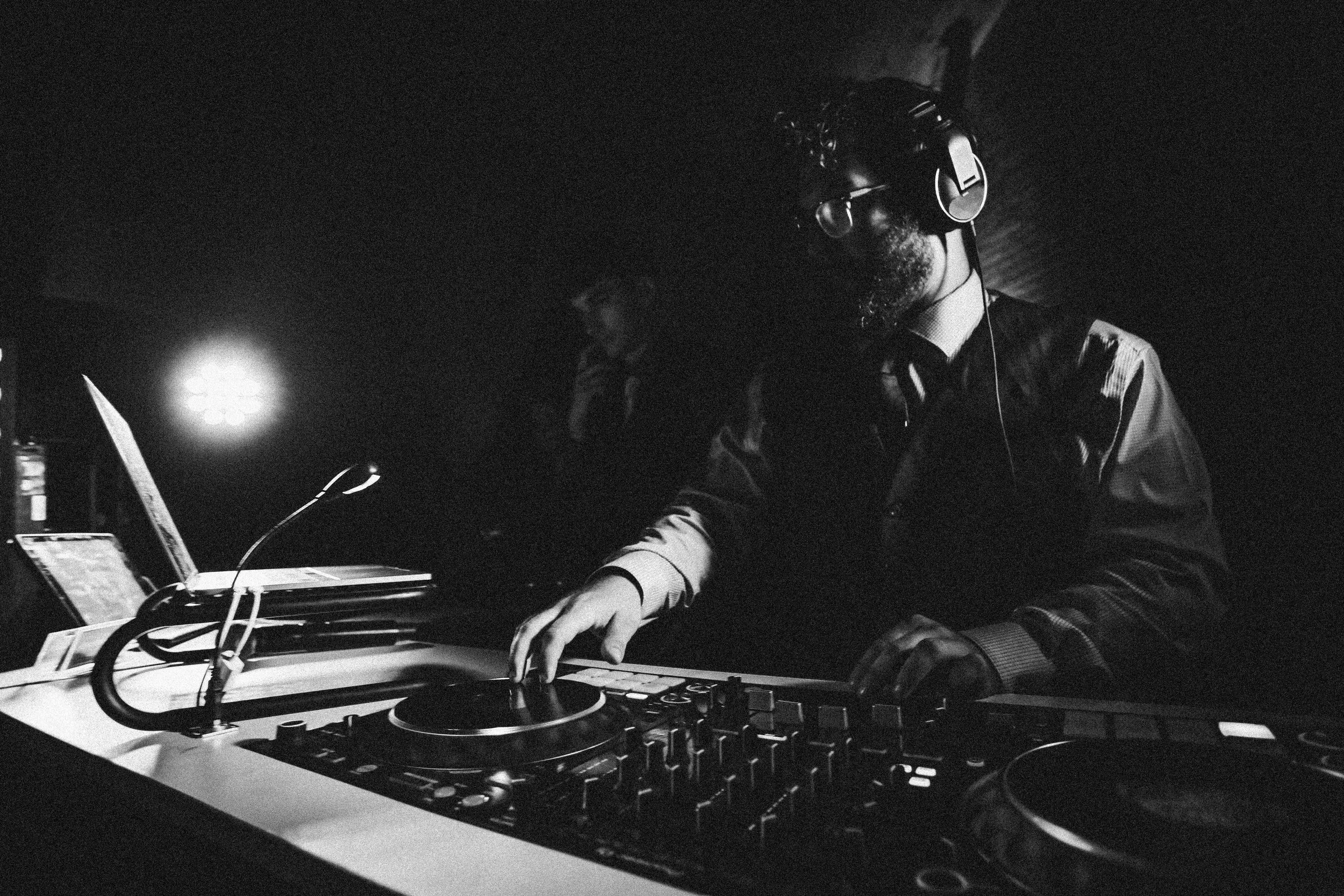 A DJ wearing headphones and sunglasses operates a DJ mixer and turntables in a dark setting, with another person in the background.