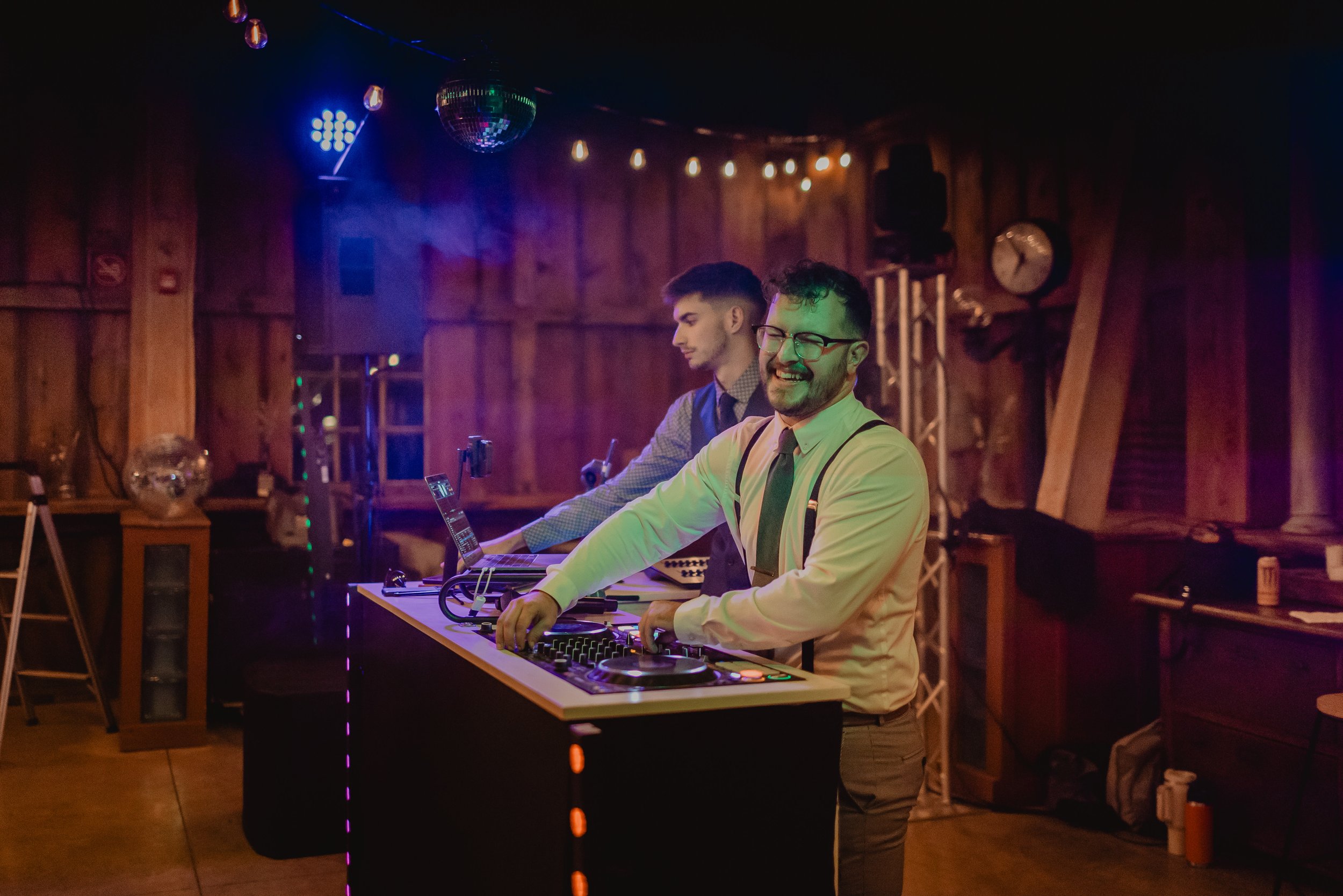 Two men dressed in formal attire DJing at a party in a wooden venue, with one smiling and the other focused on the DJ equipment.