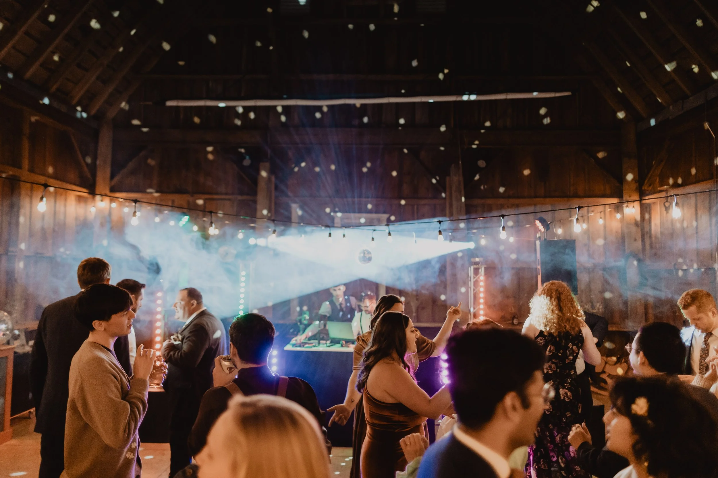 People dancing and socializing at a lively indoor party with string lights, DJ setup, and confetti in the air.