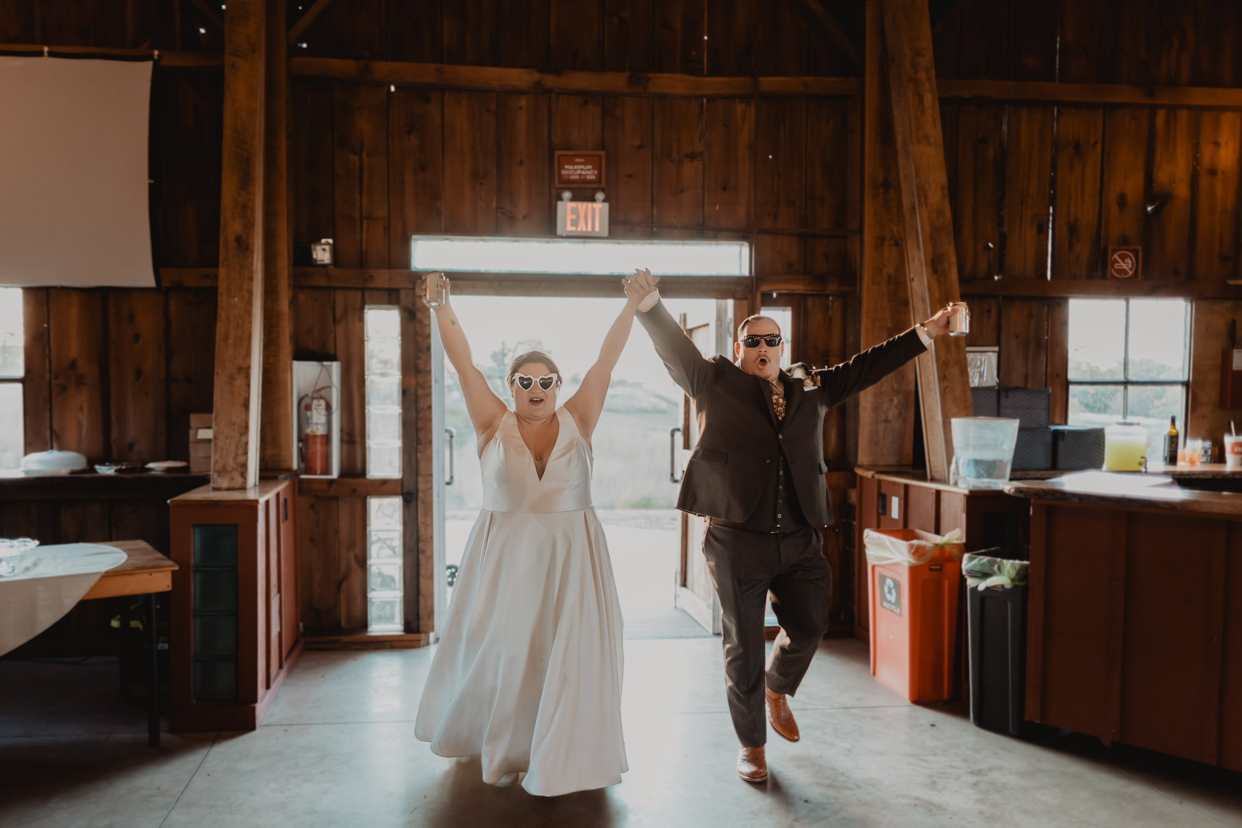 A bride and groom celebrating indoors with their arms raised in victory, both wearing sunglasses and smiling. The bride is in a white wedding dress, and the groom is in a black suit. The setting appears to be a rustic venue with wooden walls and open
