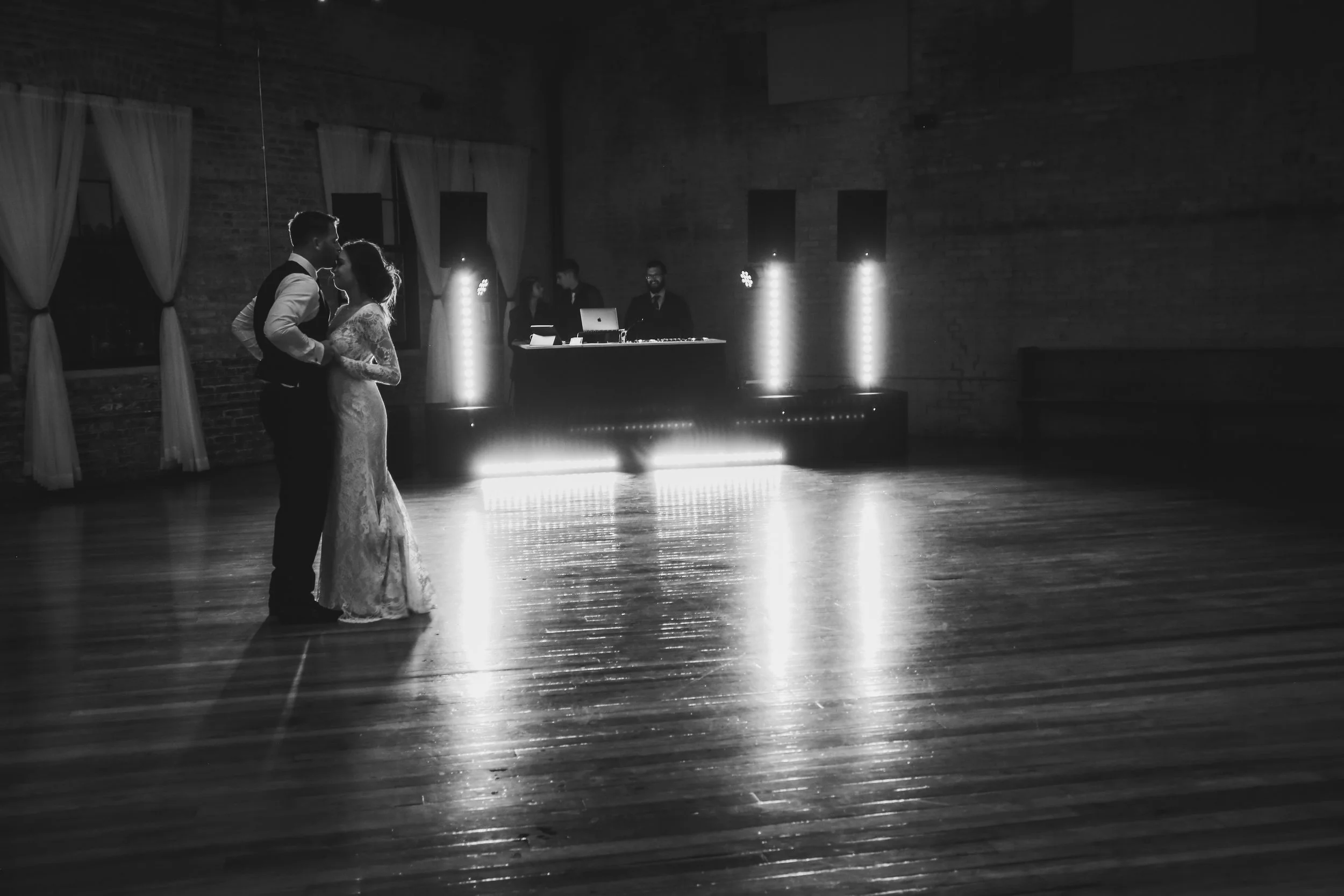 A bride and groom dancing together in a dimly lit event space with a DJ setup in the background.