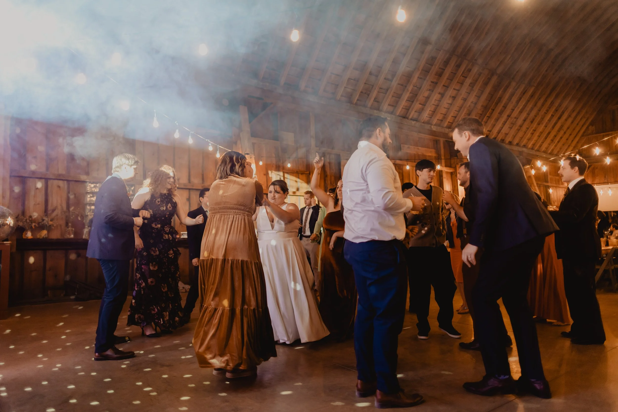 People dancing and socializing at a celebration or wedding reception in a rustic wooden barn with string lights.