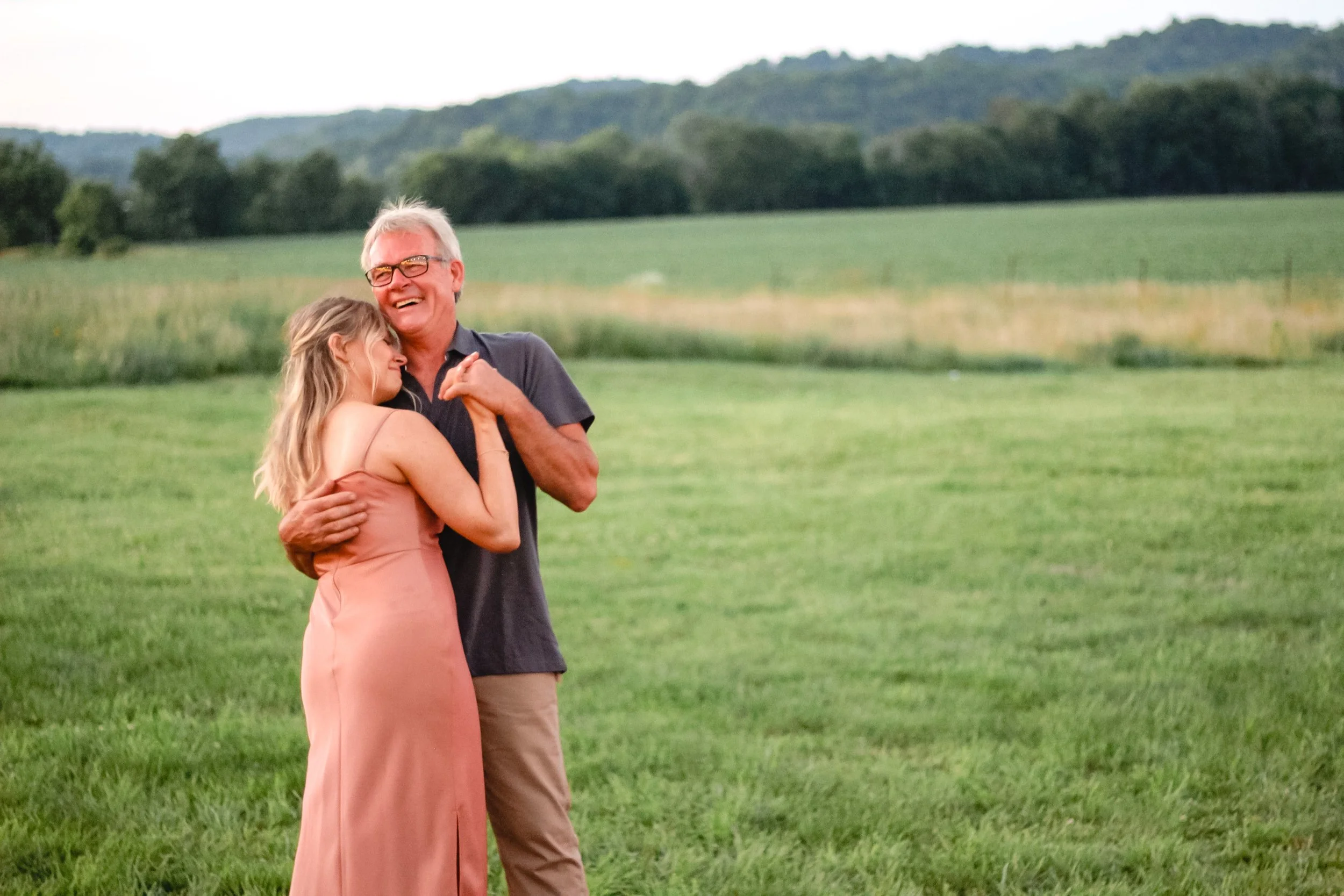 A young woman in a peach dress hugging an older man in a black shirt, both smiling, standing on a grassy field with hills and trees in the background.