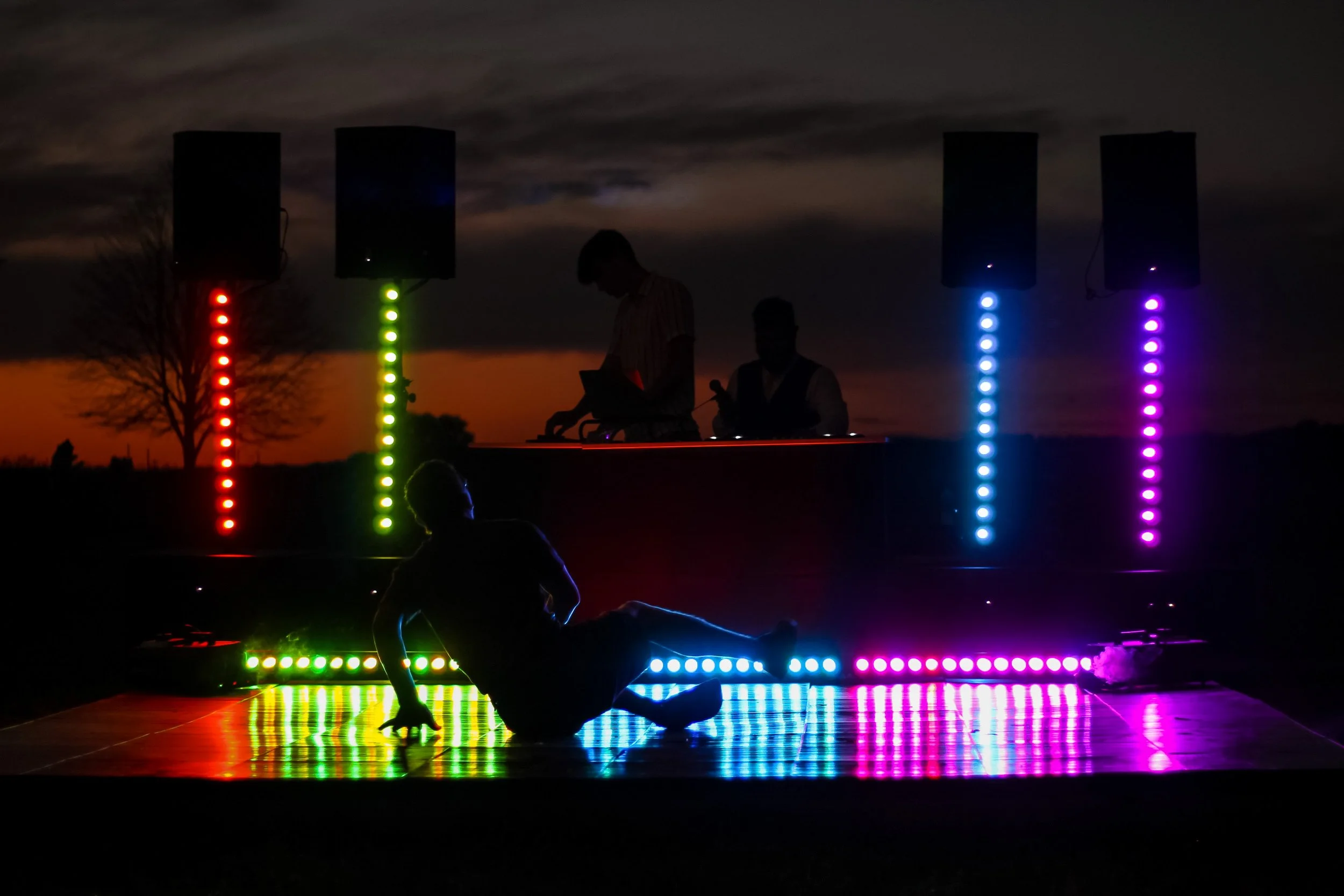 Silhouette of a person sitting on the ground dancing in front of a DJ booth with colorful LED lights, during sunset or night.