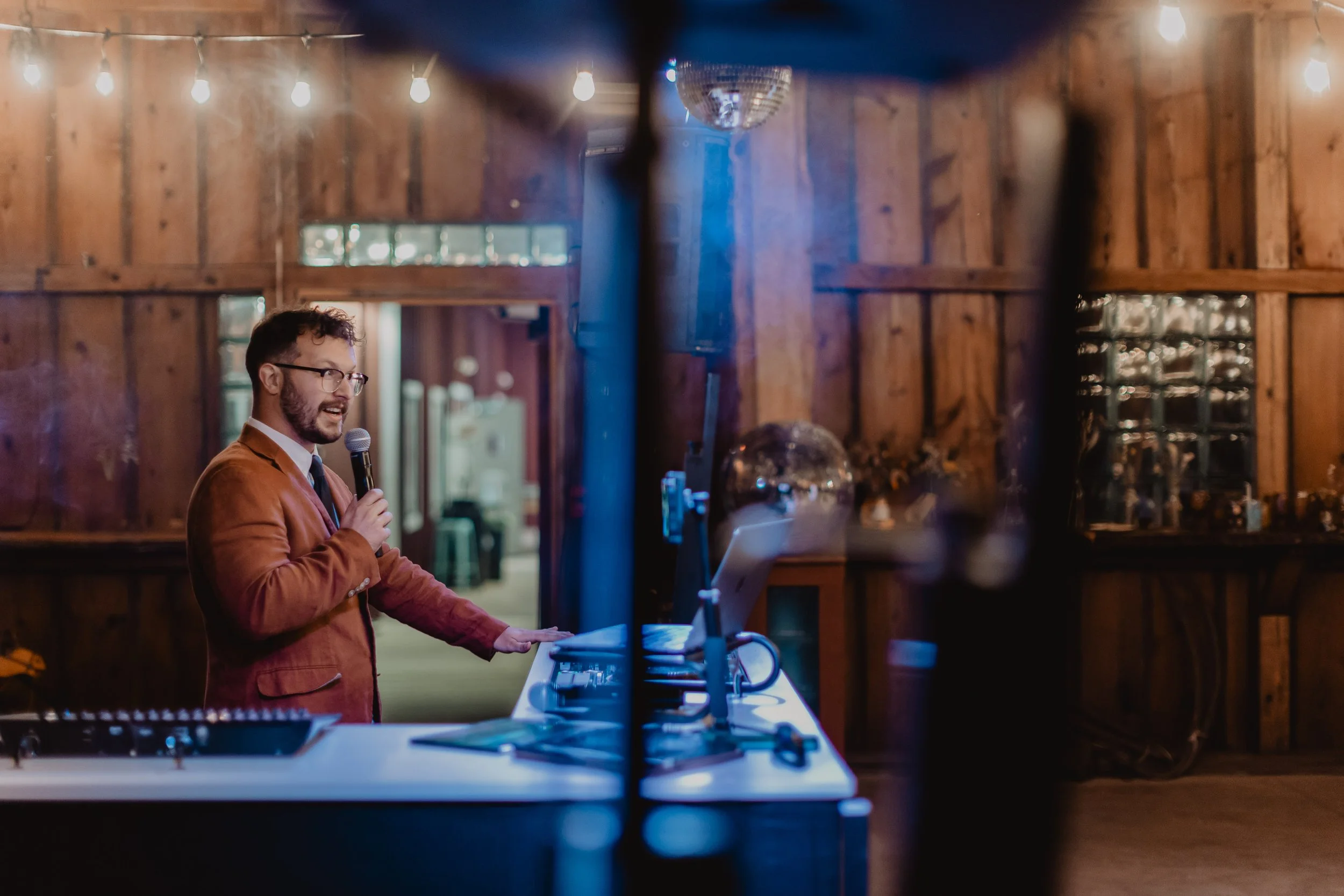 A man in a brown blazer and glasses speaking into a microphone while standing behind a DJ booth in a wooden-paneled venue decorated with string lights.