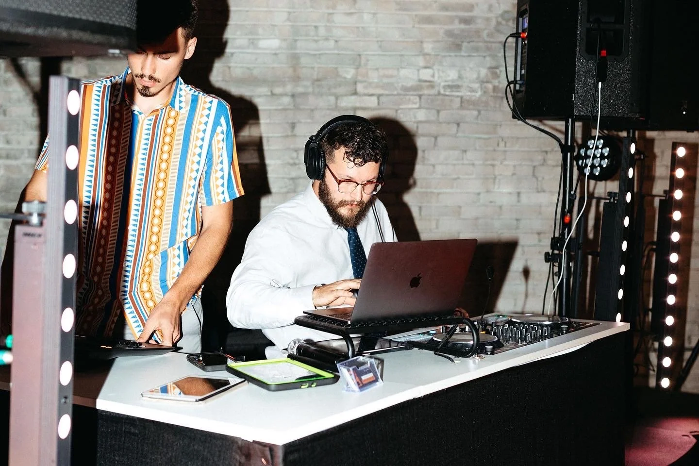Two men working at a DJ station with a laptop, audio equipment, and electronic devices in a dimly lit venue with a brick wall background.