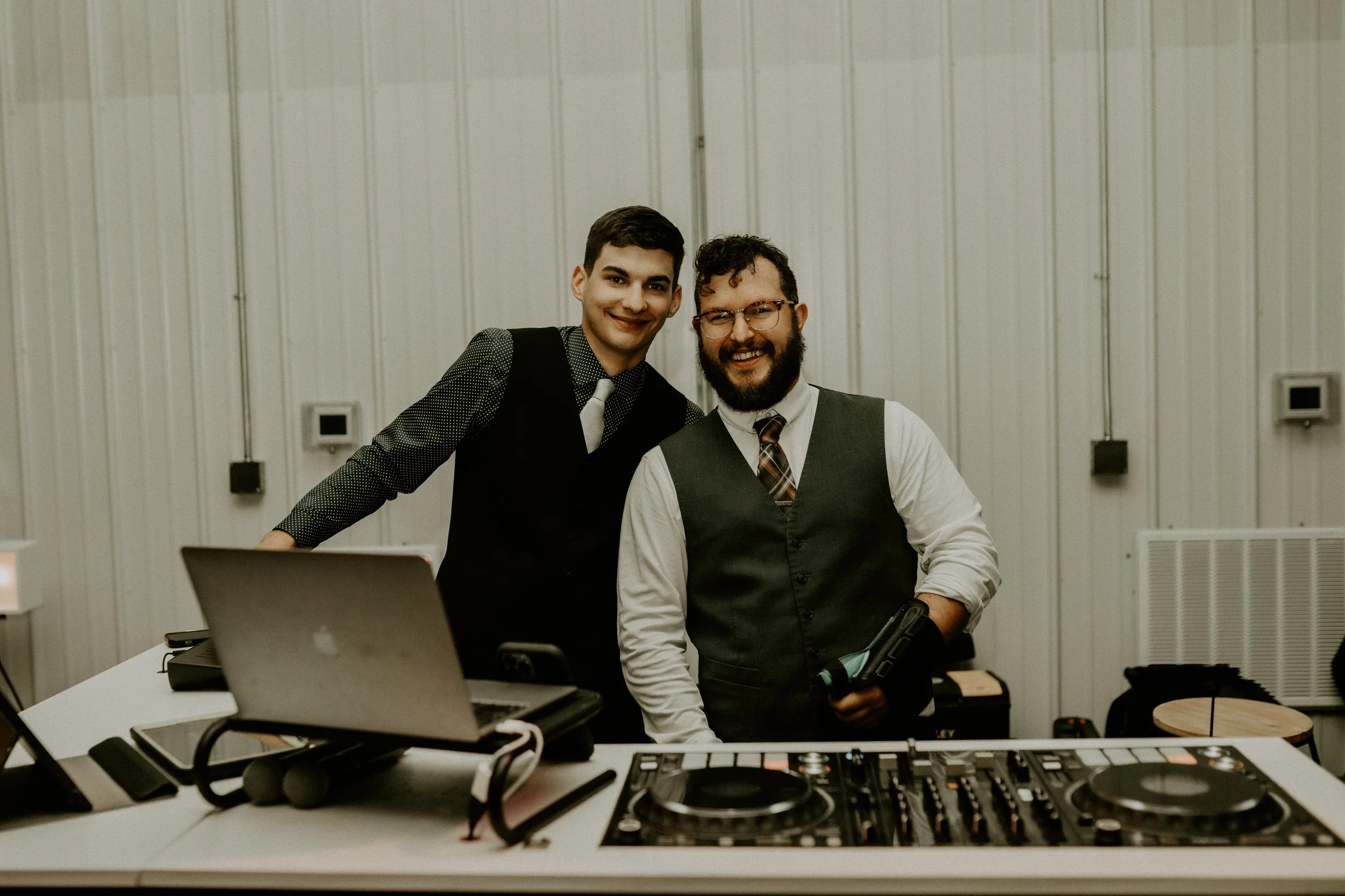 Two men smiling at a DJ setup with laptops and turntables in a room with white panel walls.