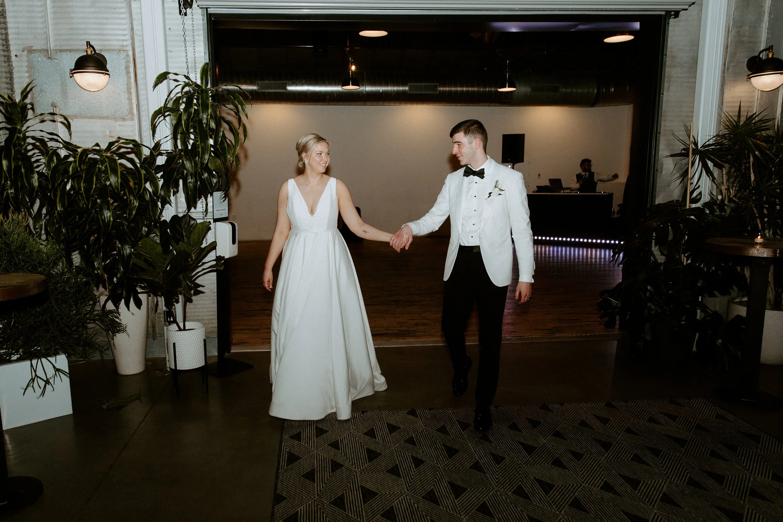 Bride and groom dancing at a wedding reception indoors, holding hands and smiling at each other, with DJ in the background.