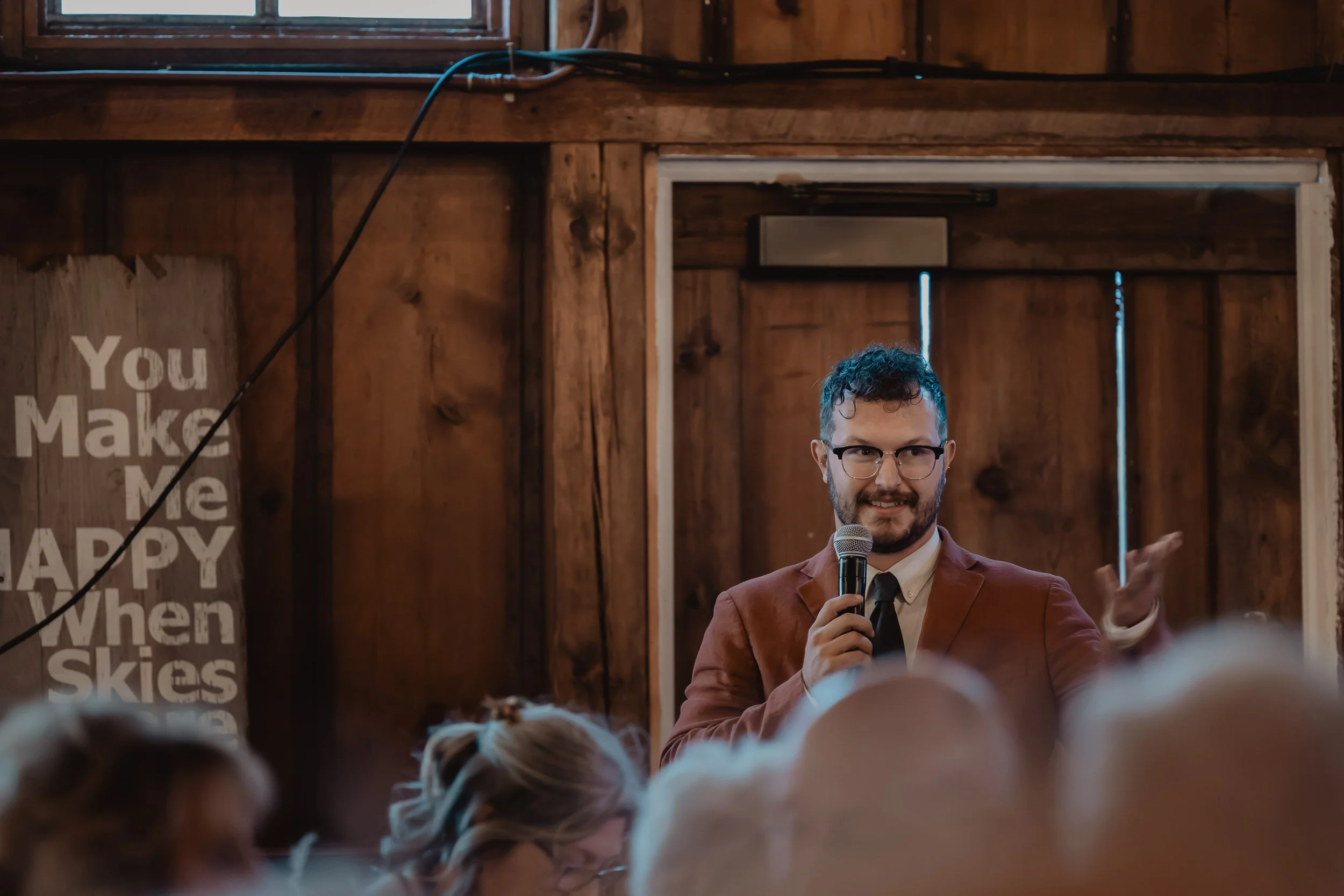 A man with glasses and a beard, wearing a brown jacket and a white shirt, is speaking into a microphone in a wooden room, with an audience in front of him.