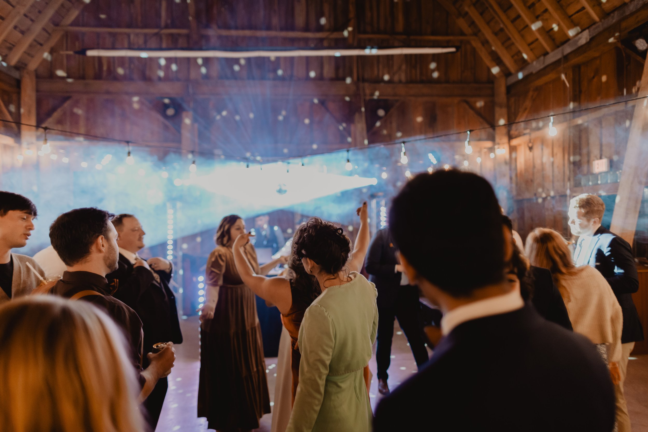 People dancing and socializing at a party or wedding reception inside a rustic wooden barn.
