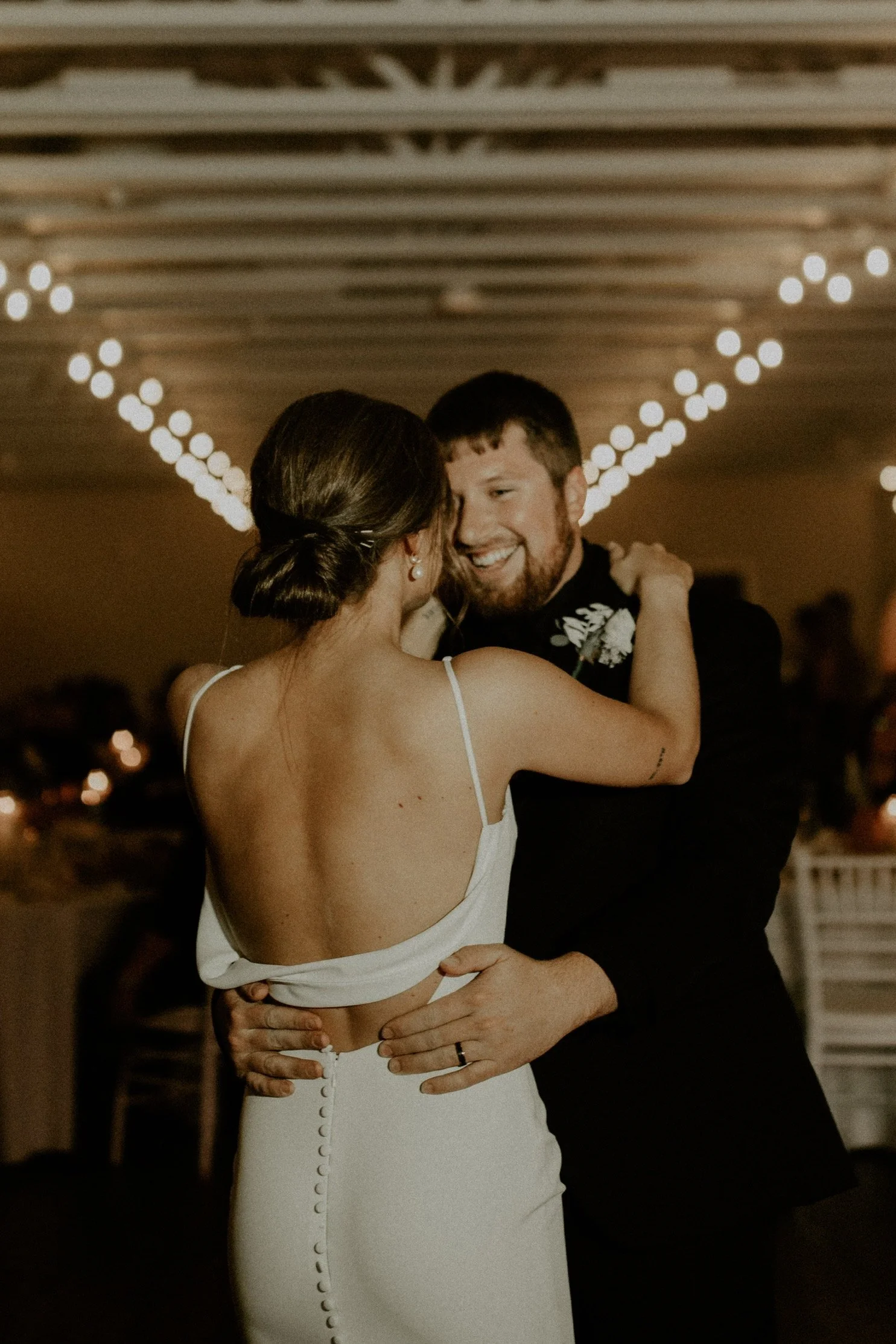 A couple is dancing closely at a wedding reception, with warm lighting and string lights above, in an indoor venue.