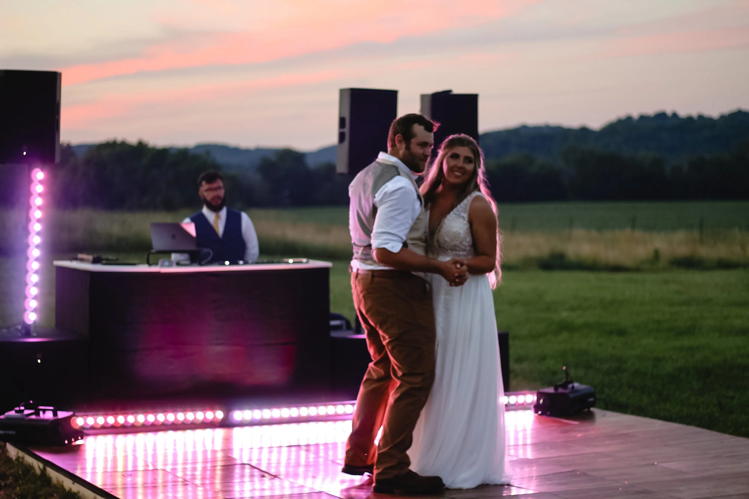 Couple dancing on outdoor wooden dance floor at sunset with DJ in background and scenic green fields.