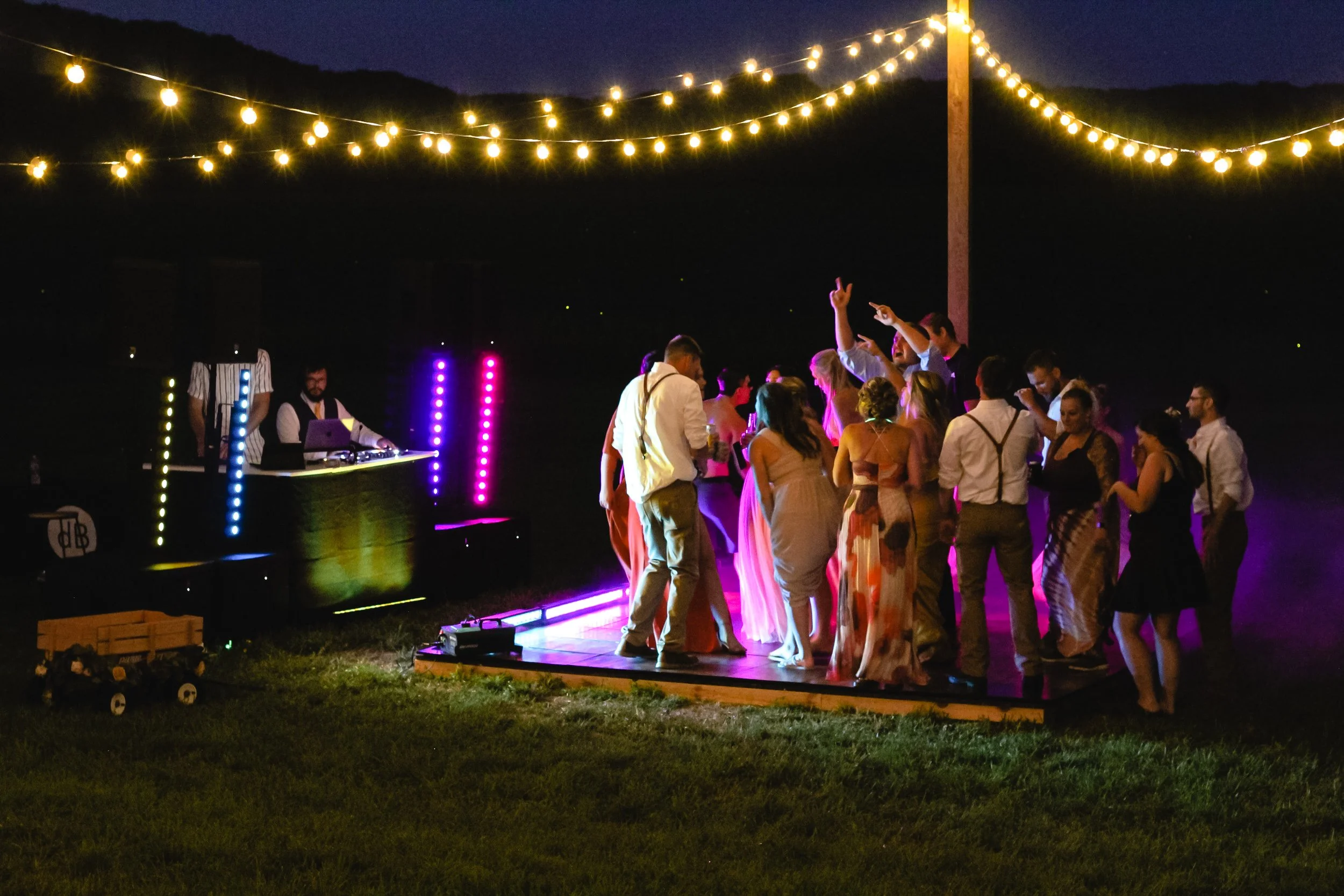 People dancing at an outdoor party at night with string lights overhead and colorful LED lights near a DJ booth.
