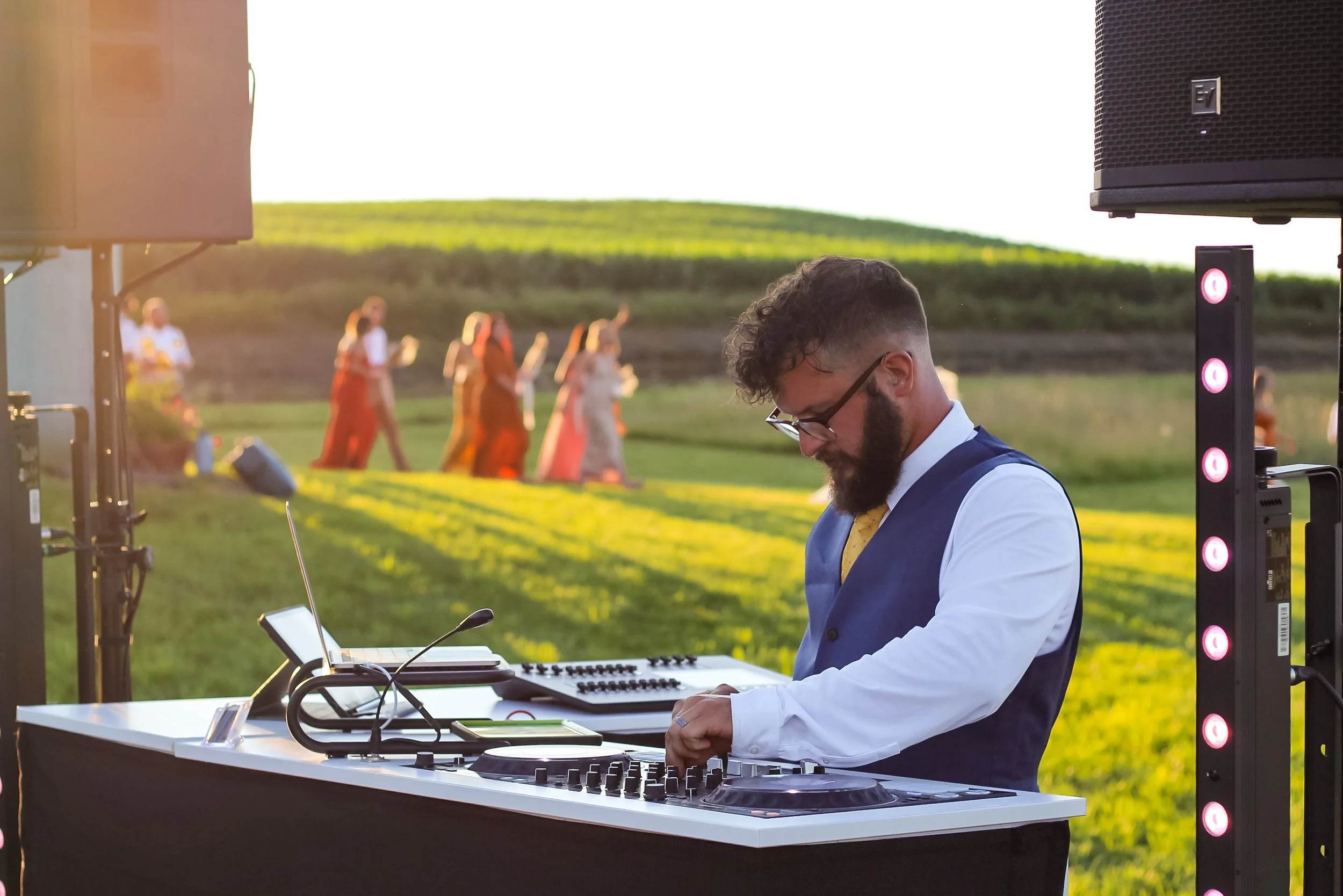 DJ wearing glasses and a vest, using a DJ controller at an outdoor event during sunset, with a group of people walking in the background on a grassy field.