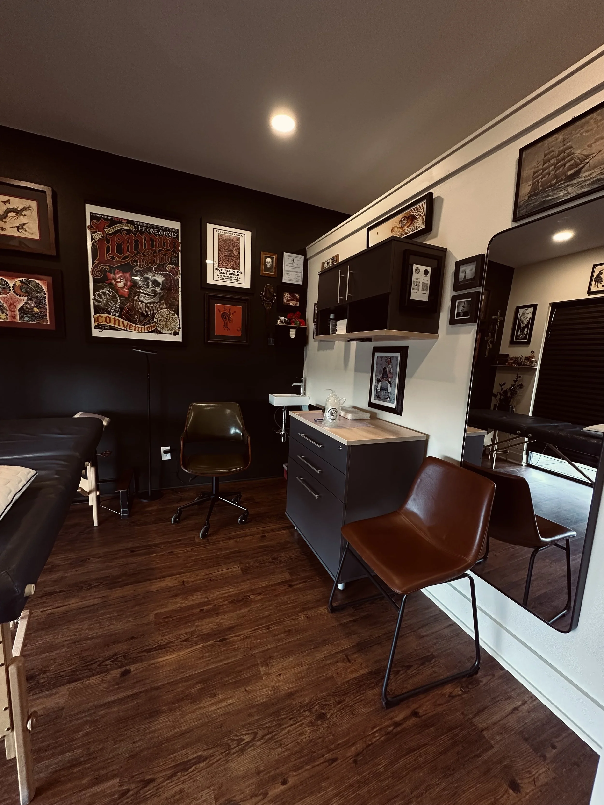 A room with framed artwork on the walls, a black cabinet with a countertop, a brown chair, a mirror, and a massage table in the corner.