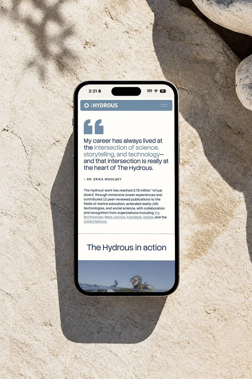 Smartphone displaying an article about The Hydrous on a sandy surface with rocks and shadows.