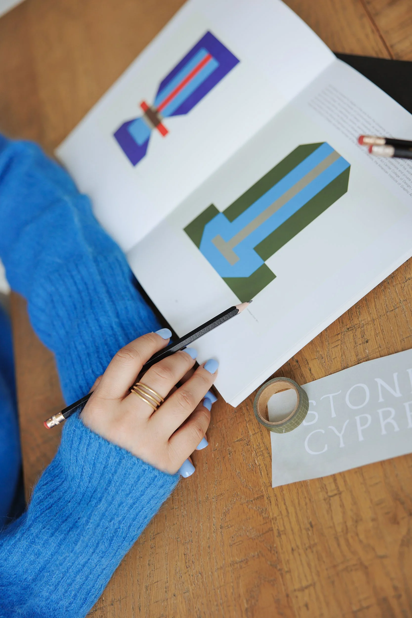 Person in blue sweater holding a pencil over a book with geometric sword designs.
