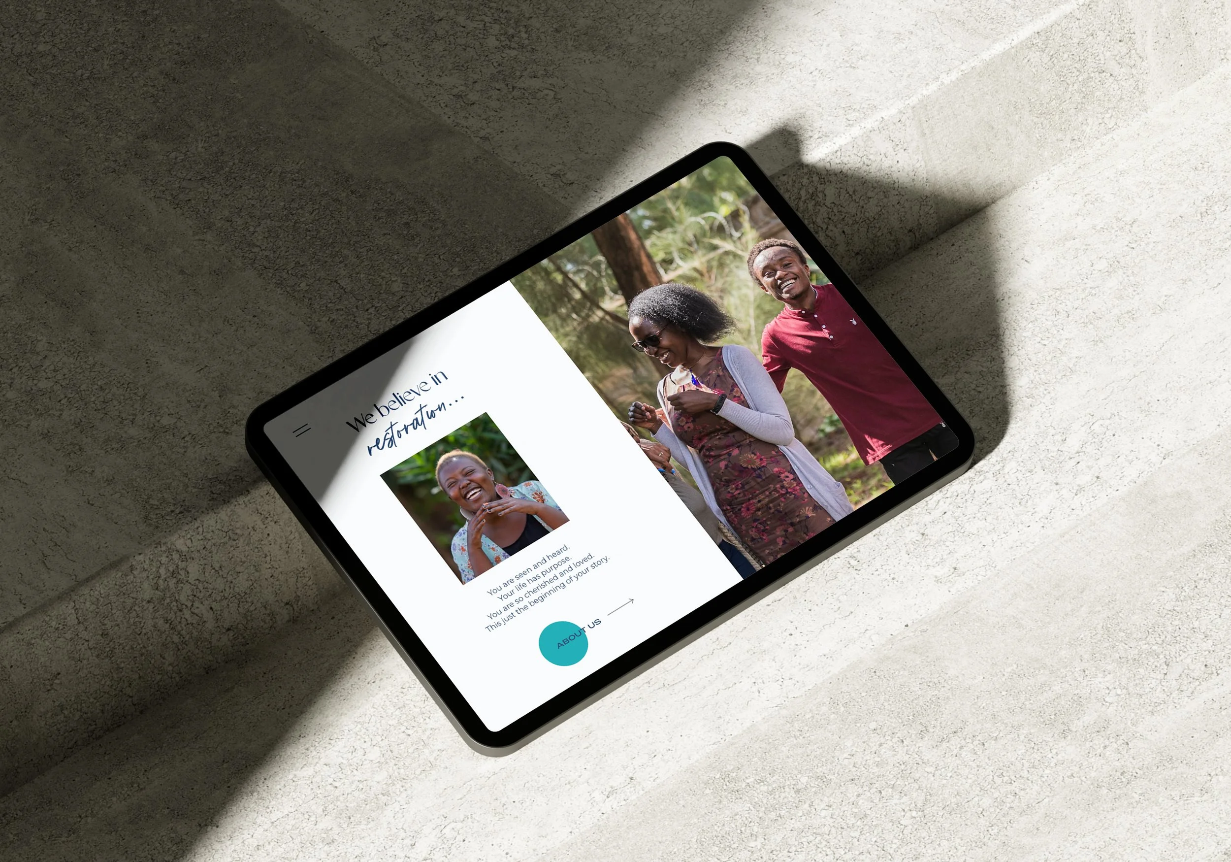 A tablet on a textured surface displaying a website with images of smiling women outdoors and text about belief in restoration.