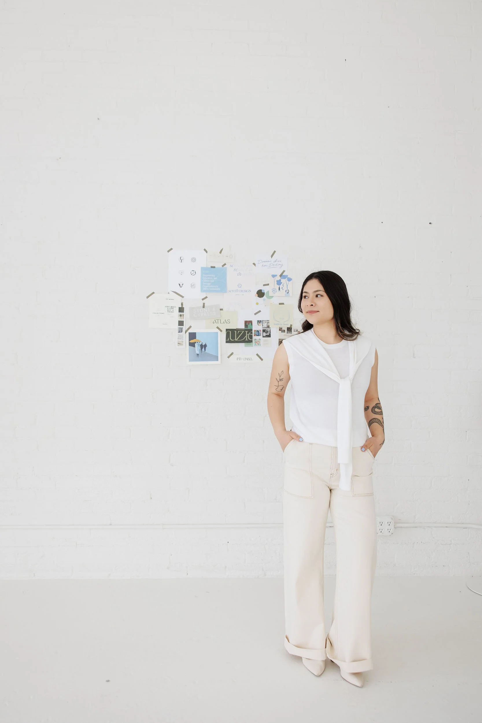 Woman in white outfit standing against a white wall with a collage of papers taped to the wall behind her.