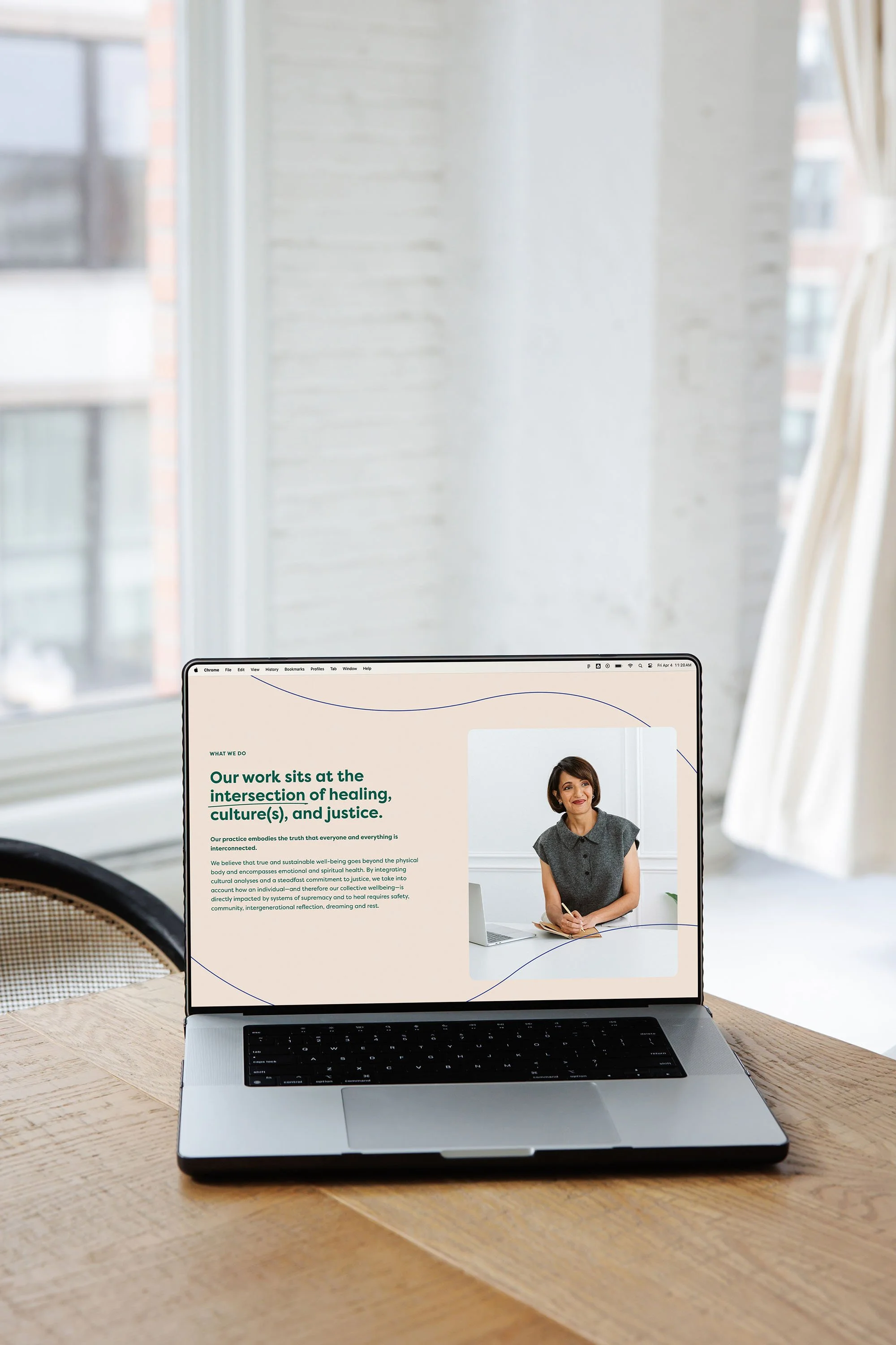 Open laptop on a wooden table displaying a webpage with a woman's photo and text about healing, culture, and justice. Bright sunlight coming through a window in a bright room.