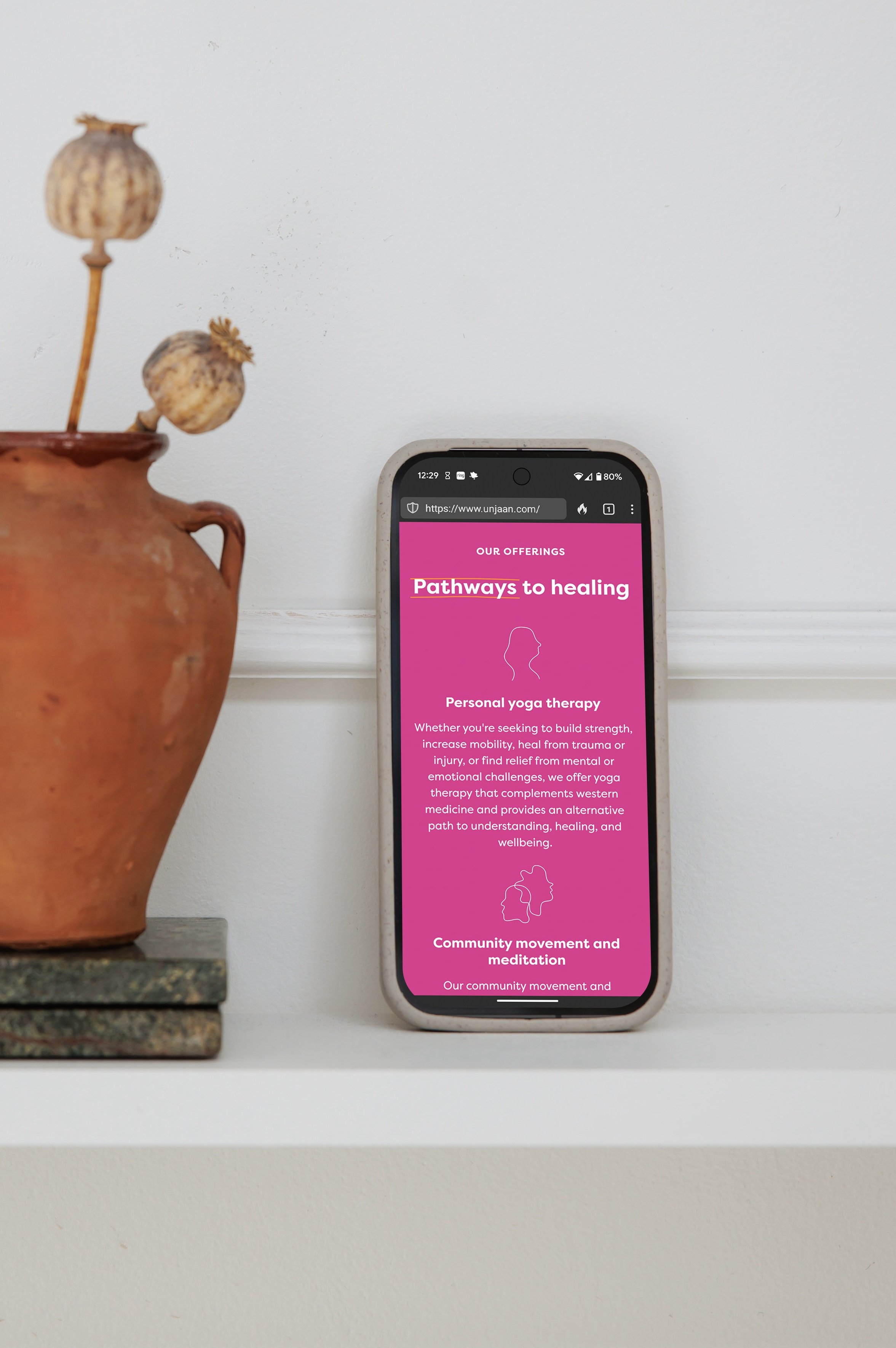 A smartphone placed on a white shelf displaying a website about healing pathways, with a terracotta vase containing dried poppy seed pods on the left side.