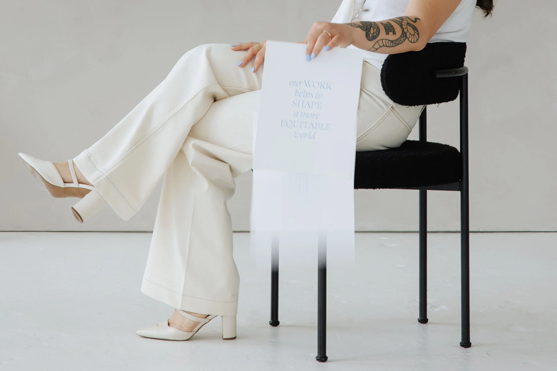 Person seated on a chair holding a paper with text 'our WORK helps to SHAPE a more EQUITABLE world'; wearing white pants and heels.
