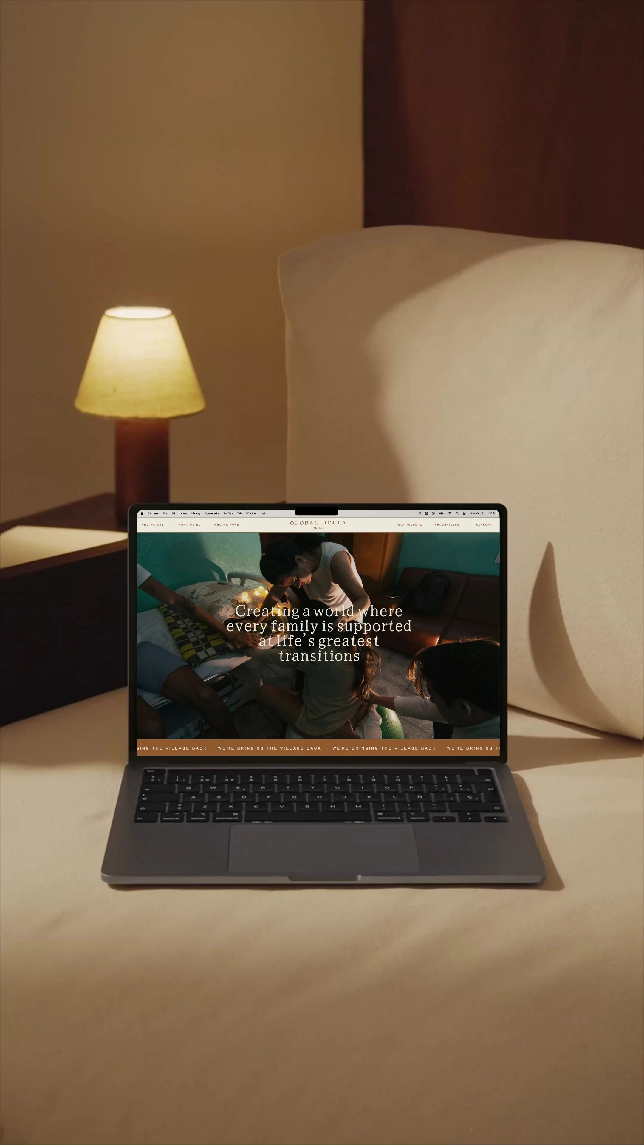 A laptop on a bed displaying a website with text "Creating a world where every family is supported at life’s greatest transitions." A lamp is lit on a bedside table.