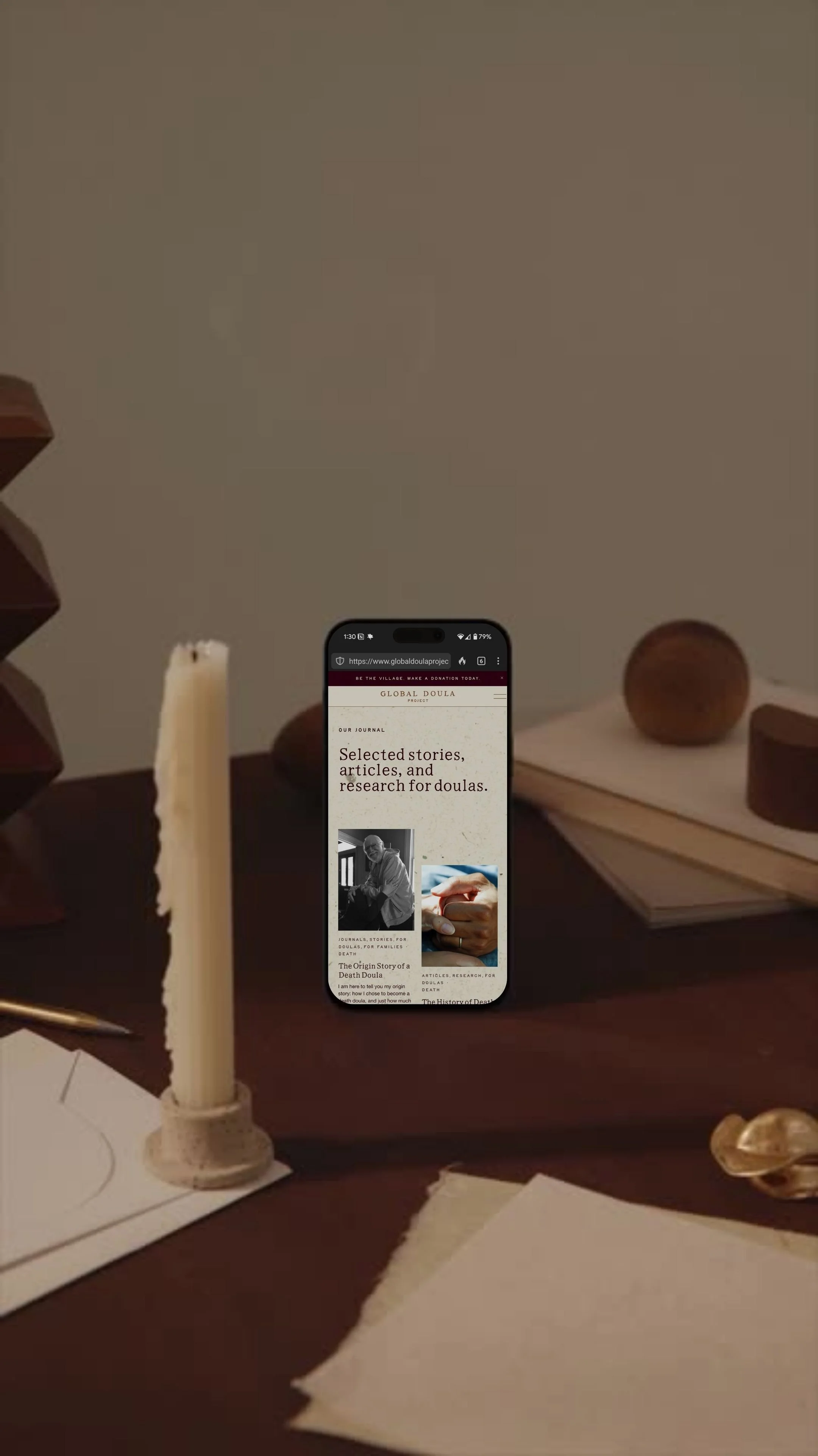 Smartphone displaying a website titled 'Global Doula' on a table with a candle, stationery, and wooden objects.
