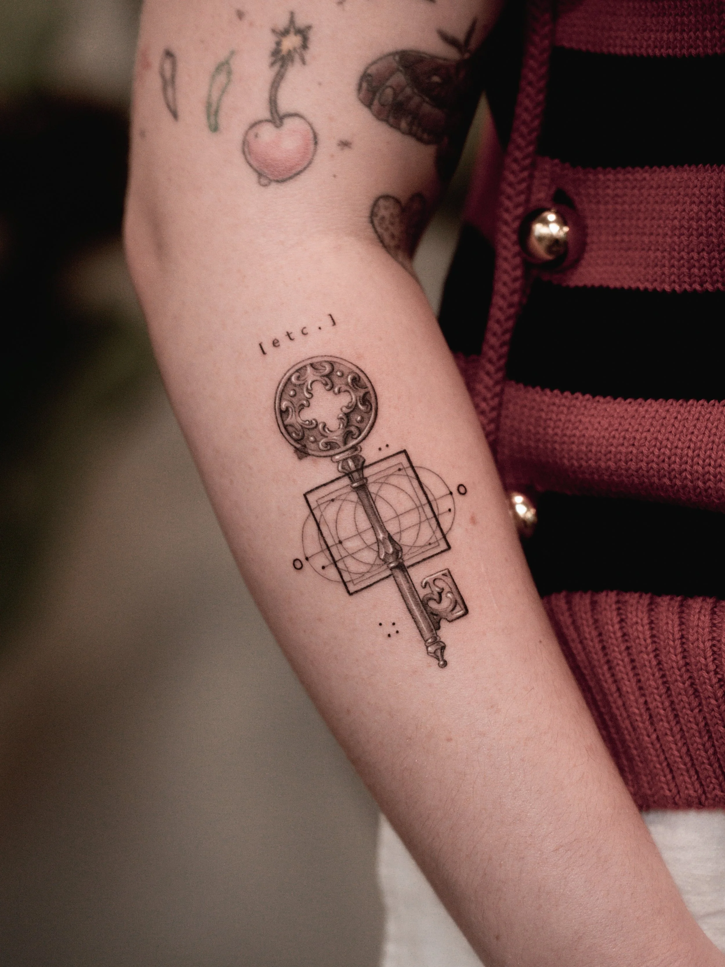 Tattoo of a key with detailed design, geometric shapes, and the text 'letc:)' on a person's arm with additional tattoos.