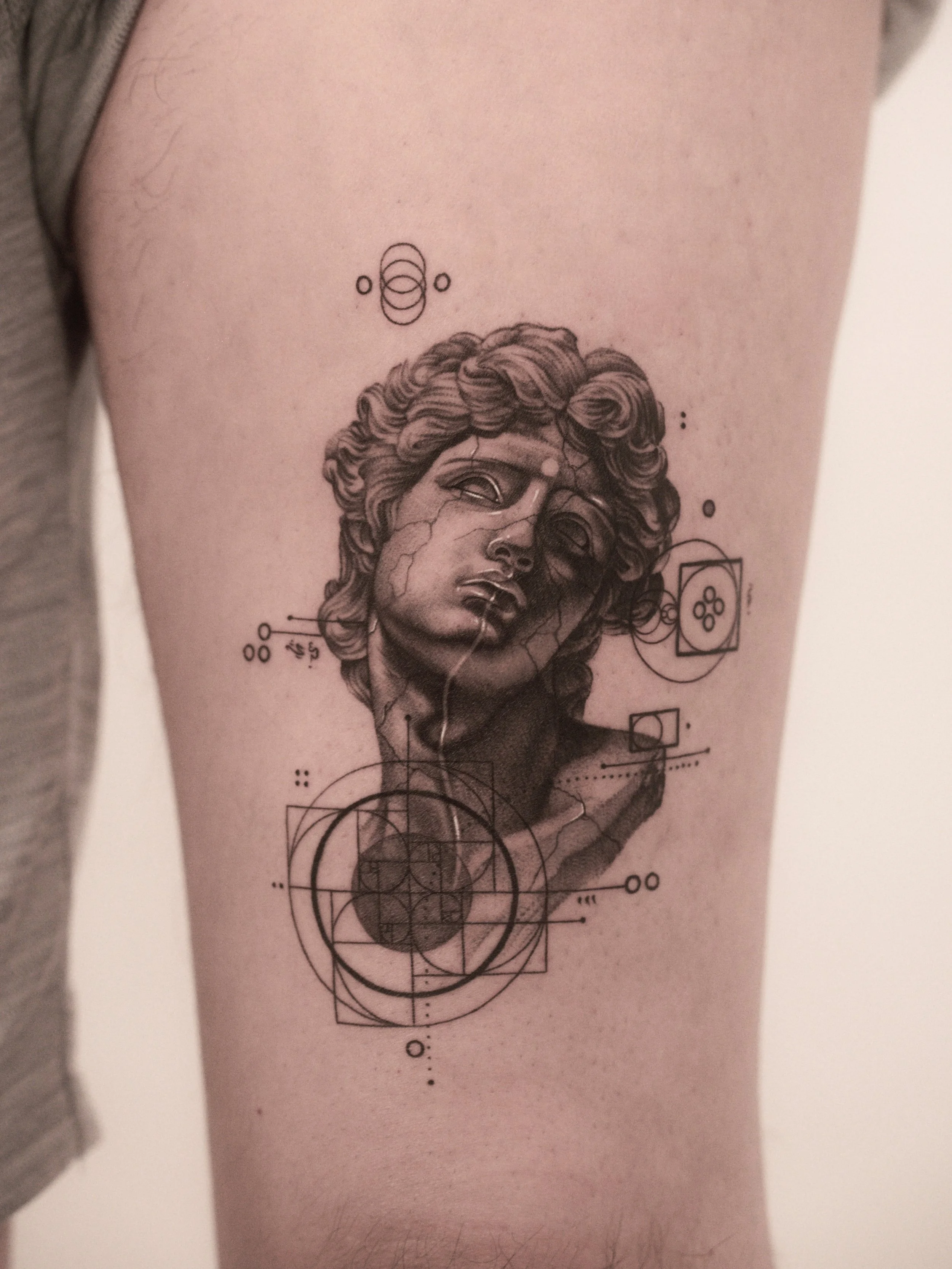 Tattoo of a classical Greek sculpture head surrounded by geometric and scientific symbols on forearm.