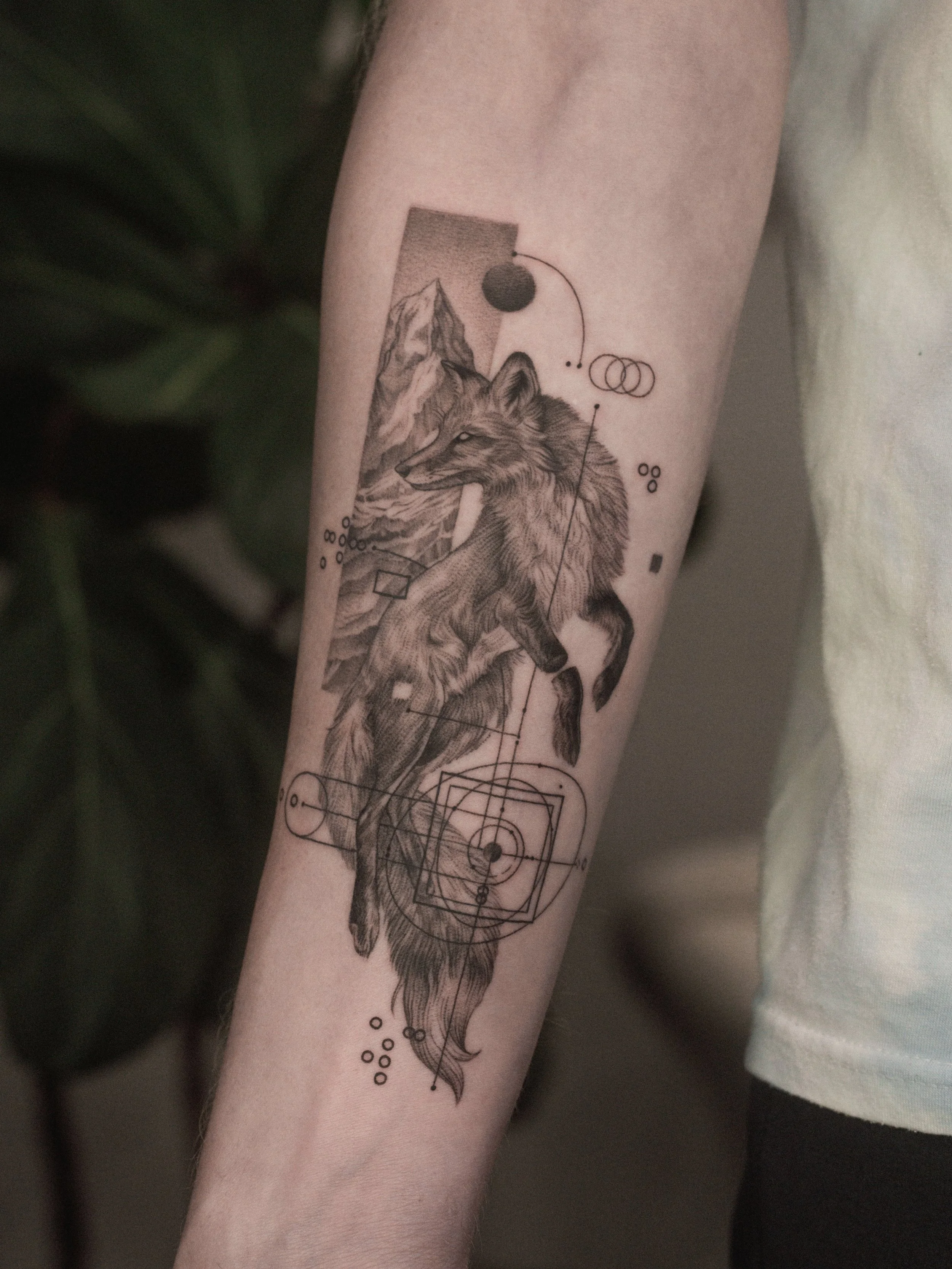 Detailed black and gray tattoo of a wolf with mountain scenery, geometric patterns, and celestial symbols on a person's forearm.