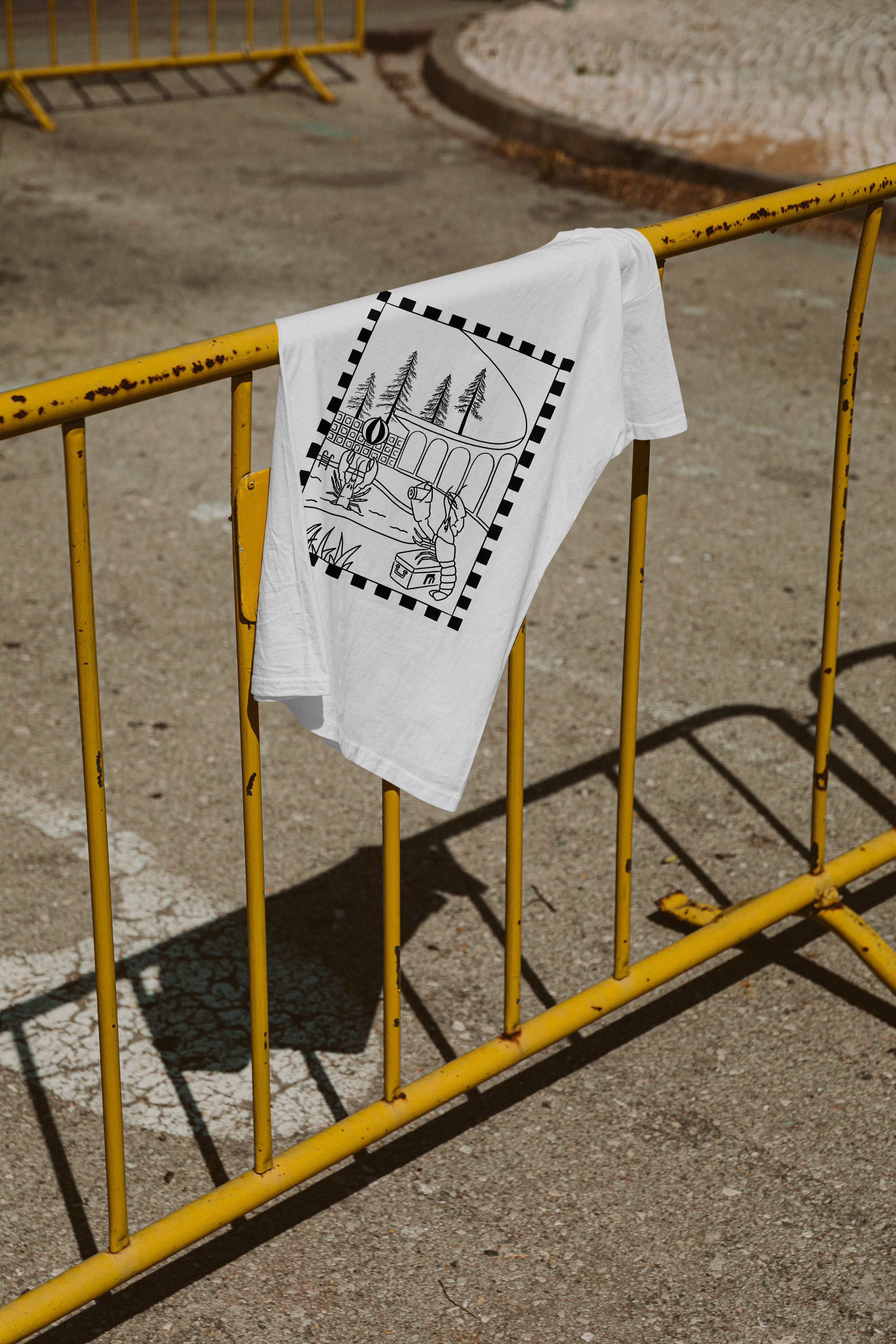 A white T-shirt with a black line drawing of a landscape featuring tall trees, a bridge, and a person fishing, hanging on a yellow metal barricade, outdoors on a sunny day.