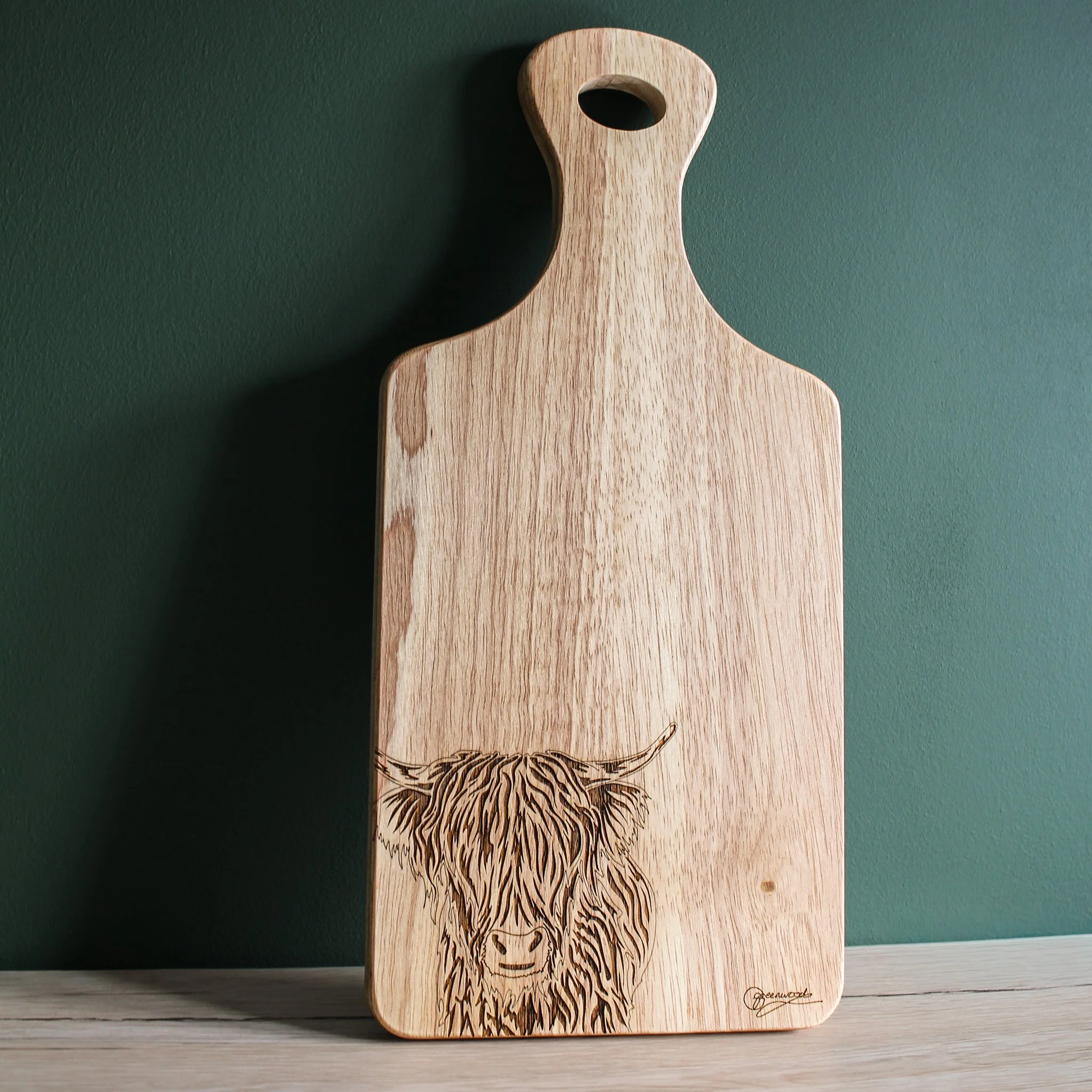Paddle shaped wooden chopping board engraved with hand drawn highland cow, displayed upright against a dark green wall.