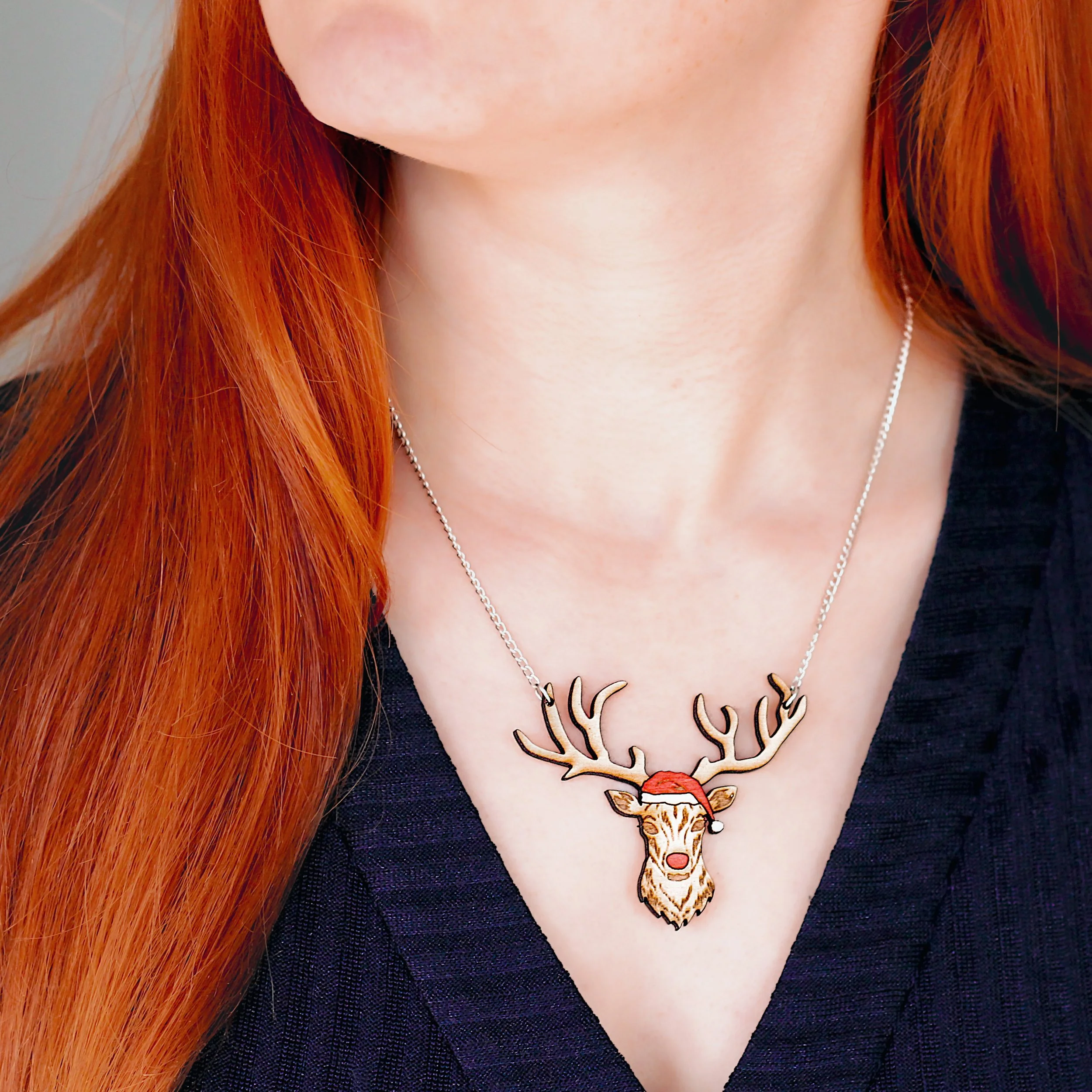 Woman wearing Rudolph the red nose reindeer necklace which has a large, wooden Rudolph pendant. It is attached to a delicate silver chain on either side of its antlers.