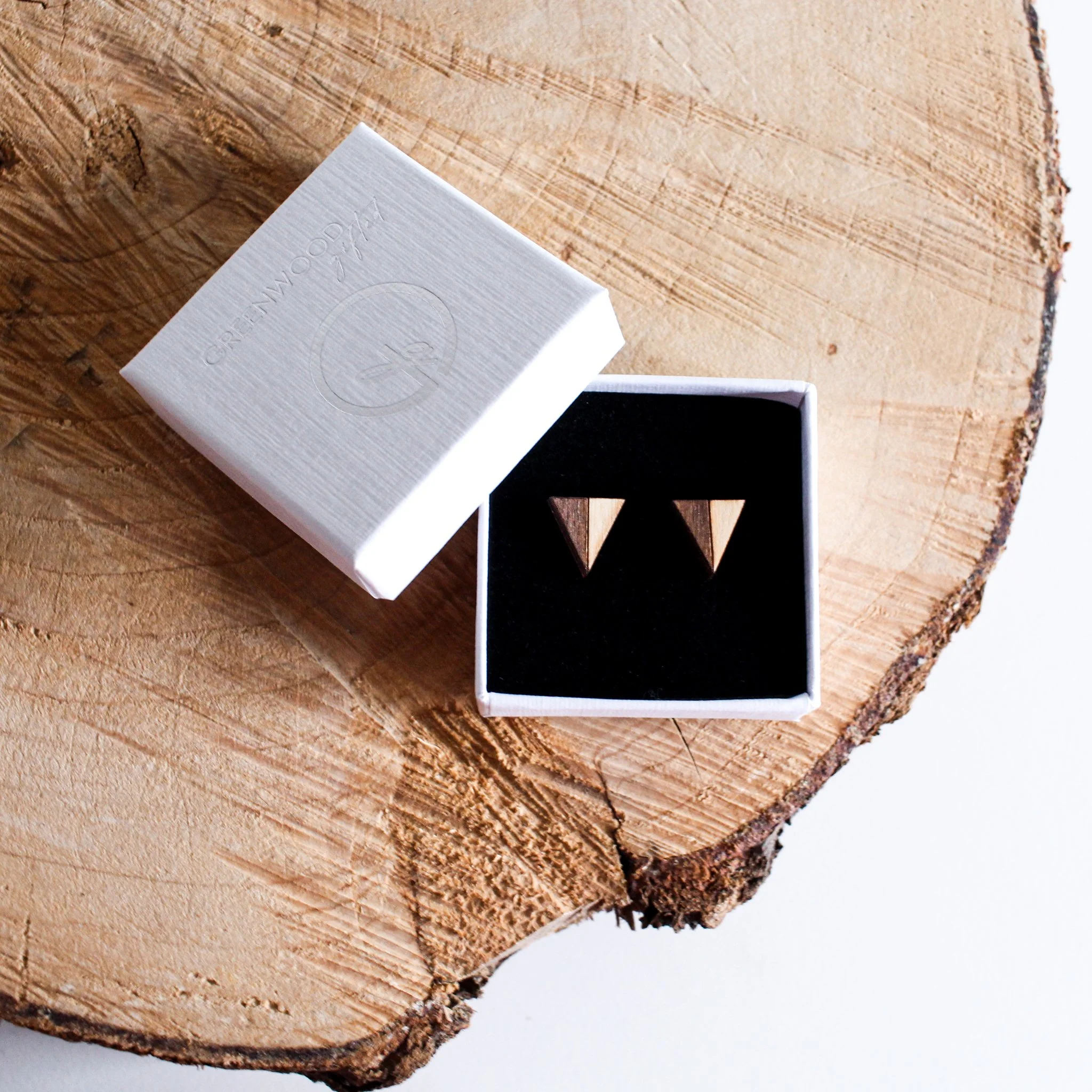 Triangle stud wooden earrings in a gift box. The earrings sit inside the box on a black foam pillow. The lid is open and to the side.