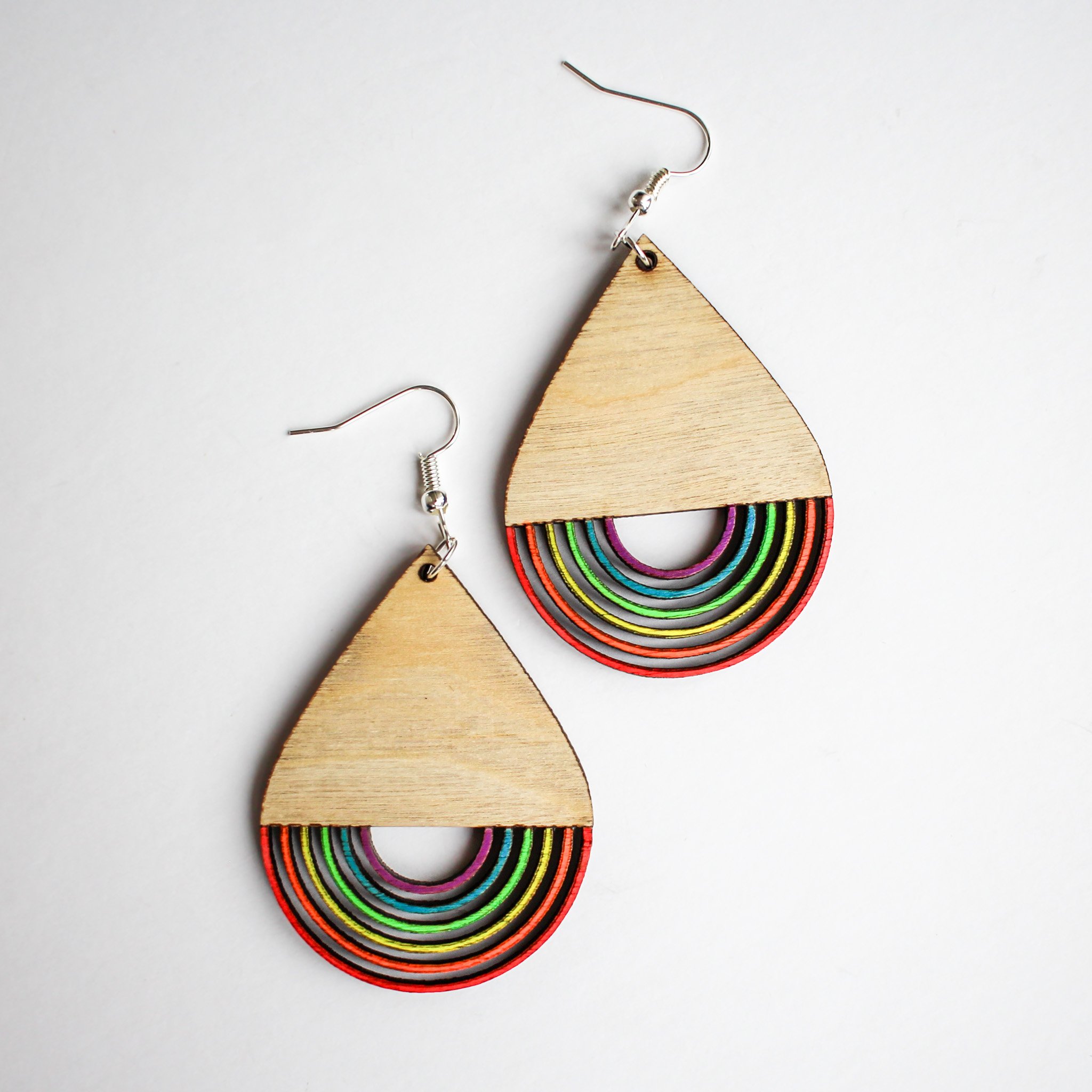 Teardrop wooden earrings with a cutout, upside down rainbow design at the base of the teardrop. The wood is birch wood and light in colour with hand painted fluorescent accents on the rainbow.