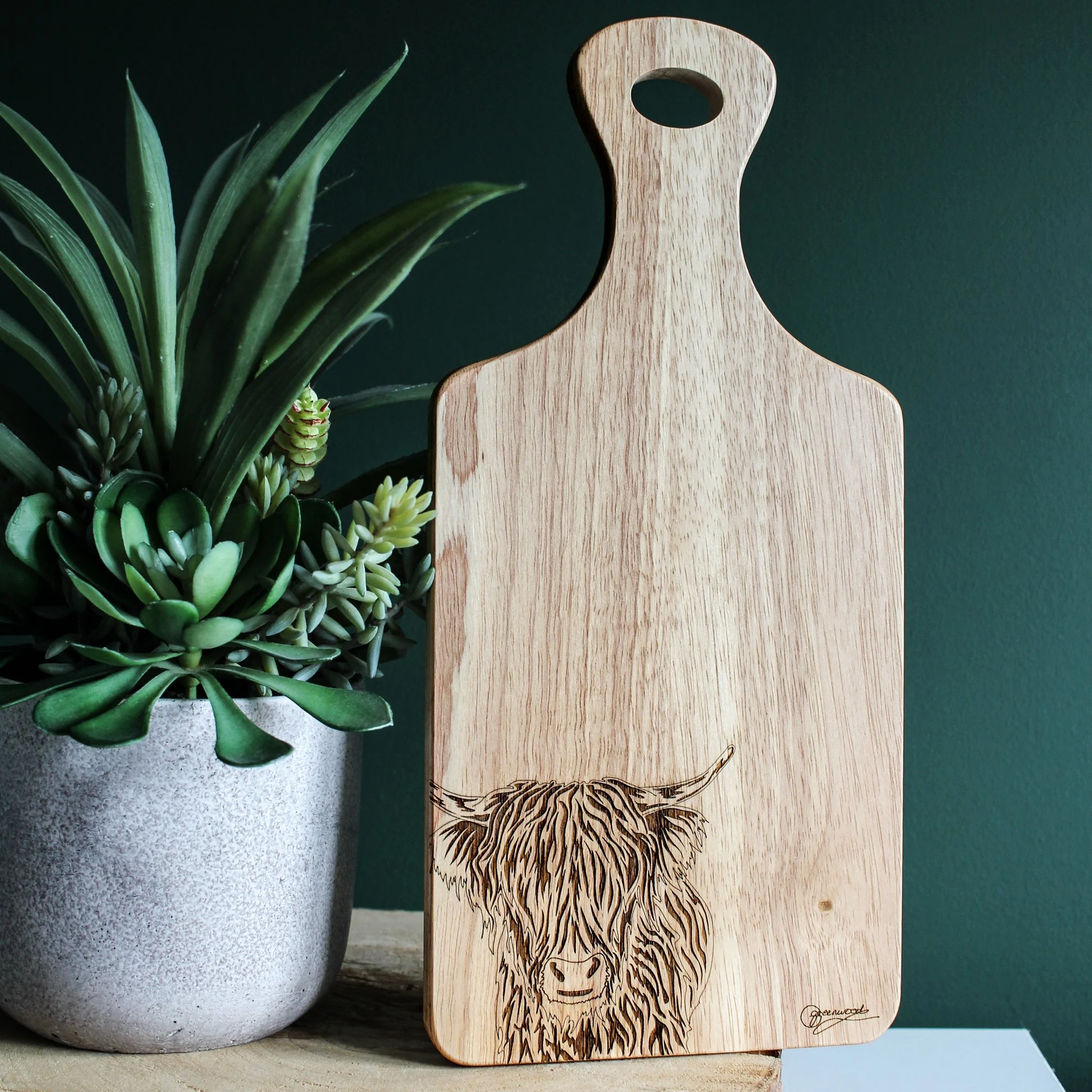 Highland cow chopping board displayed next to a plant against a dark green wall. The board is made from hevea wood and engraved with original art and the artists signature.
