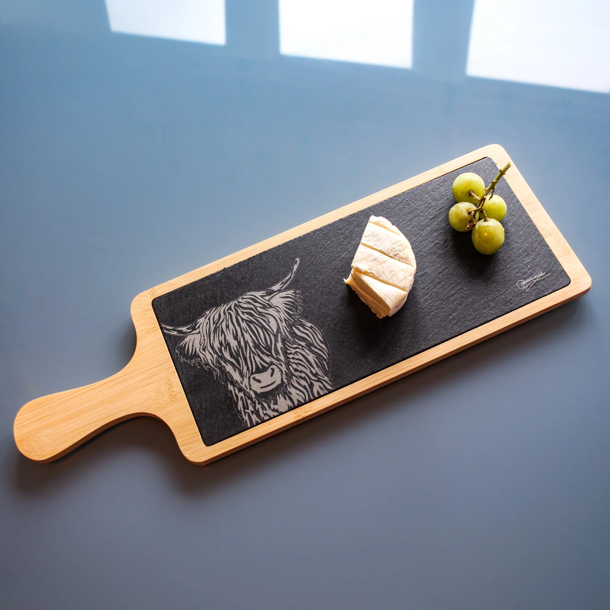 Bamboo and slate serving platter with highland cow drawing engraved on the slate. Shown in use with slate insert inside bamboo tray, cheese and grapes on top.