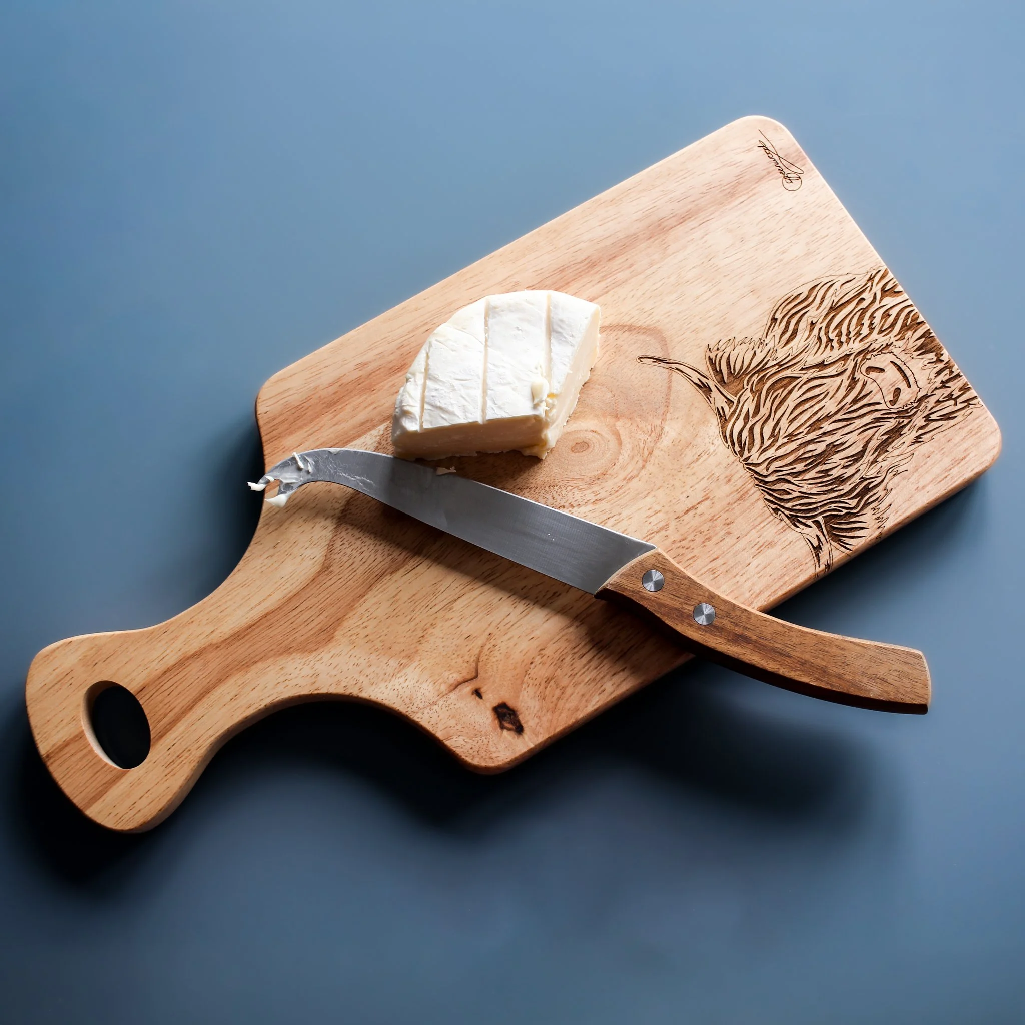 Lifestyle shot of paddle shaped chopping board made from wood, displayed with cheese and knife on top. The board is engraved with an original highland cow design and the artist signature.