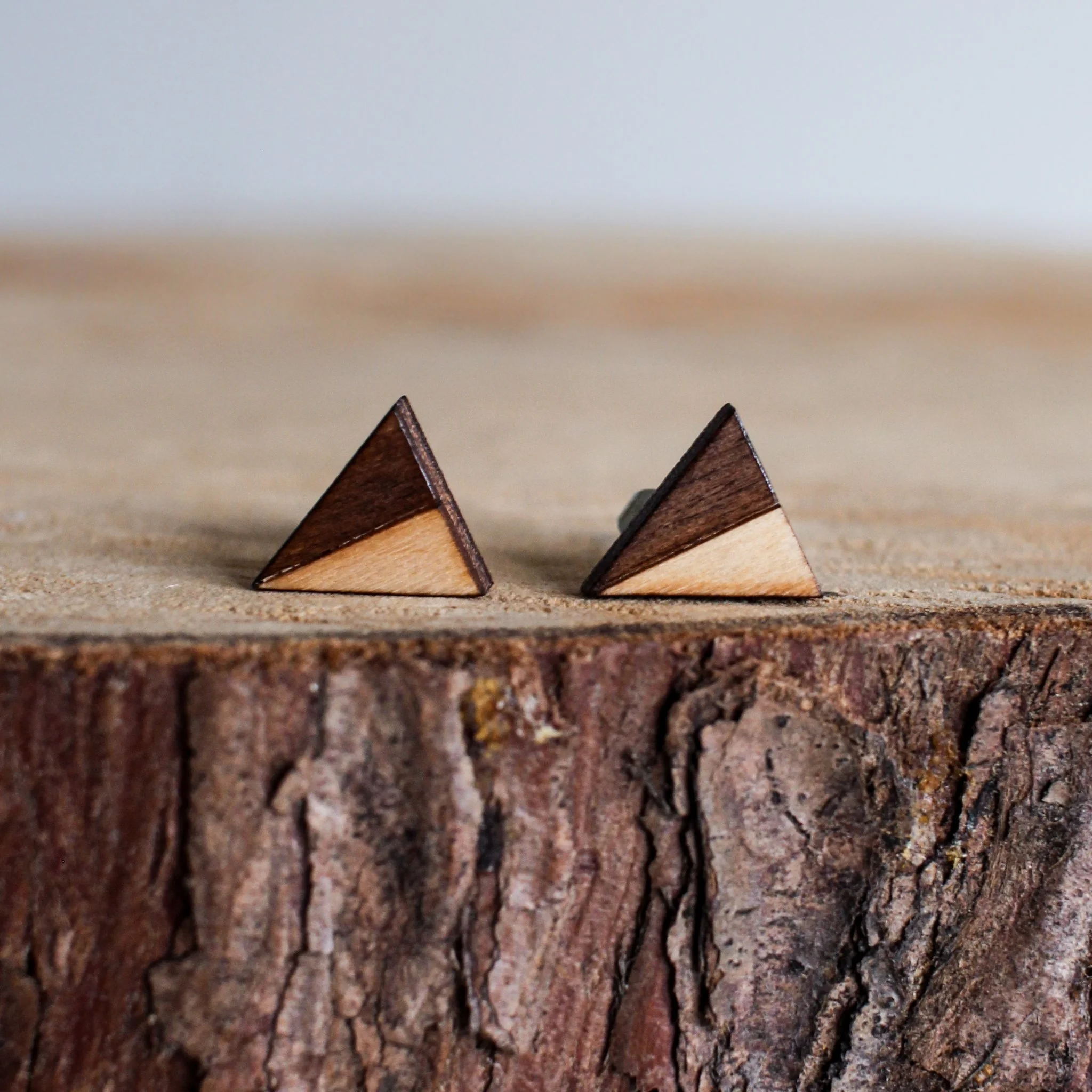 A pair of wooden studs on a log slice. The stud earrings are two toned. Half on each is dark, rich brown like bark and the other half is a pale, golden blonde.